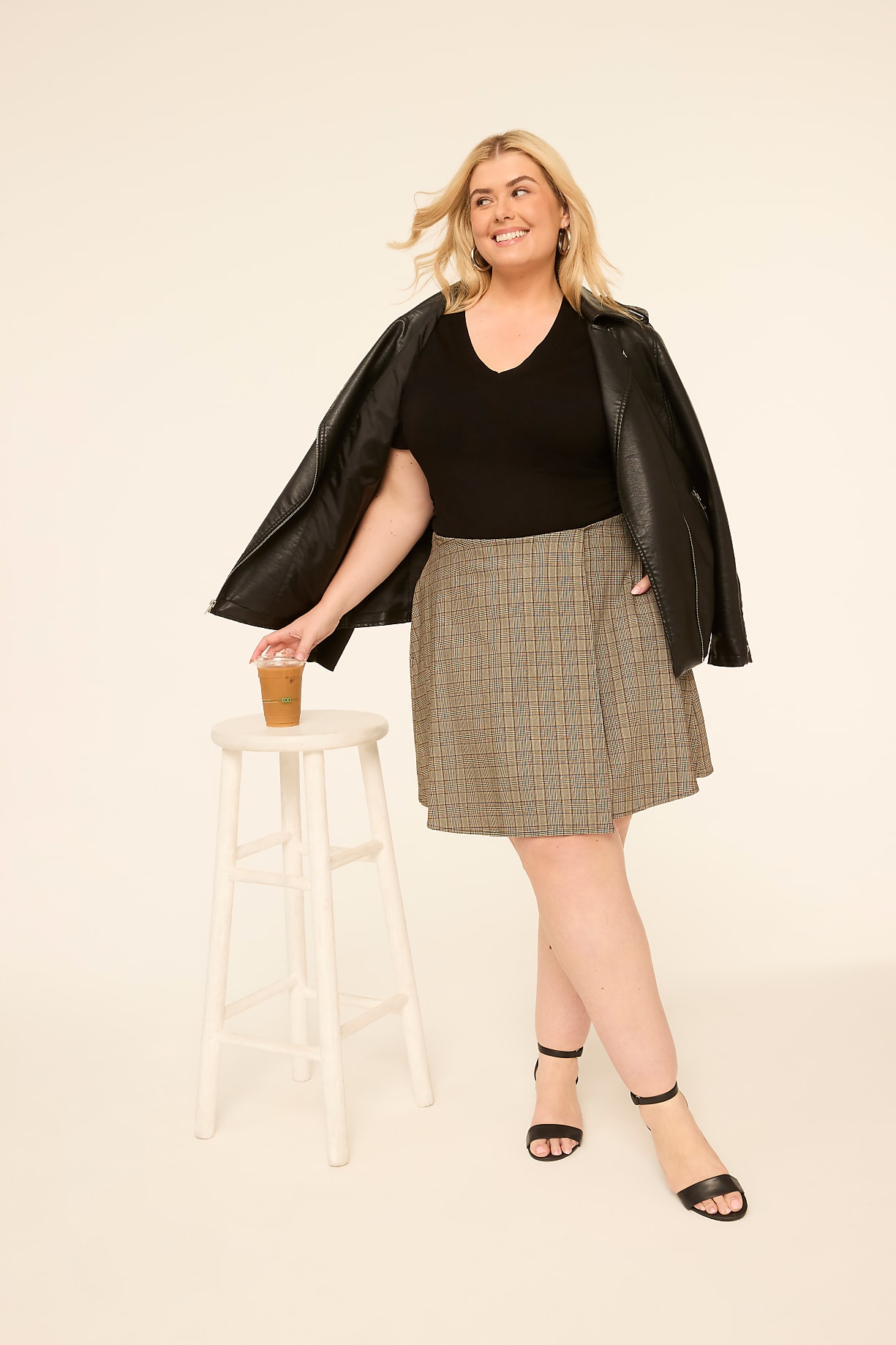 A woman in a black top, leather jacket, and plaid skirt stands next to a stool with a coffee cup, against a plain, light-colored background. Product Image 5