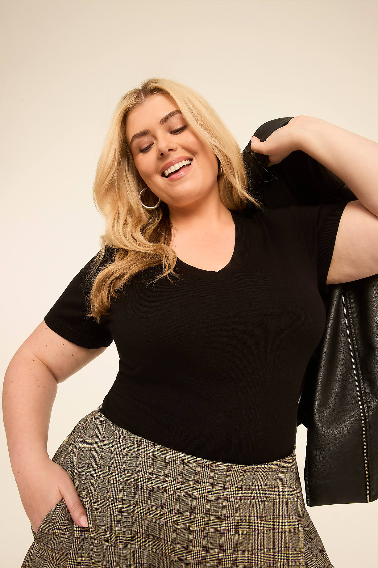 A smiling woman in a black v-neck shirt and plaid skirt poses with a leather jacket draped over her shoulder against a plain, light-colored background. Product Image 9