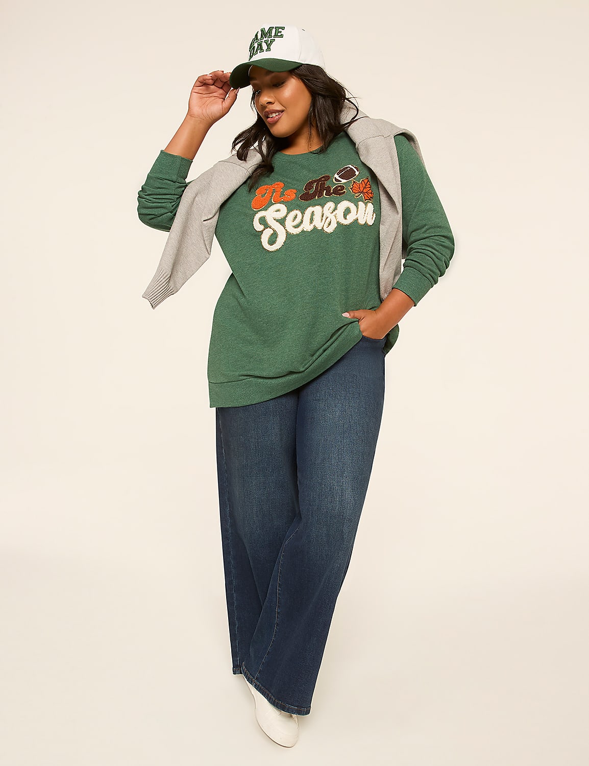 A woman in a green sweatshirt and dark jeans poses against a plain, light-colored background, holding the brim of her "Game Day" hat. Product Image 3