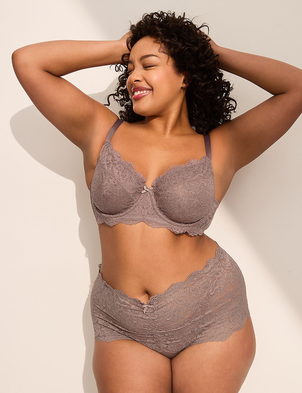 A smiling woman in a lace bra and shorts poses with her hands behind her head against a plain, shadowed background. Product Image 4