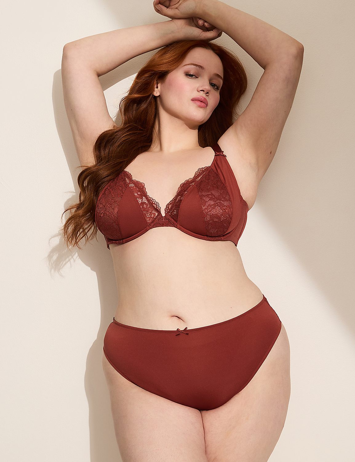 A woman with auburn hair poses in a rust-colored lace bra and matching underwear against a plain, light-colored wall. Product Image 4