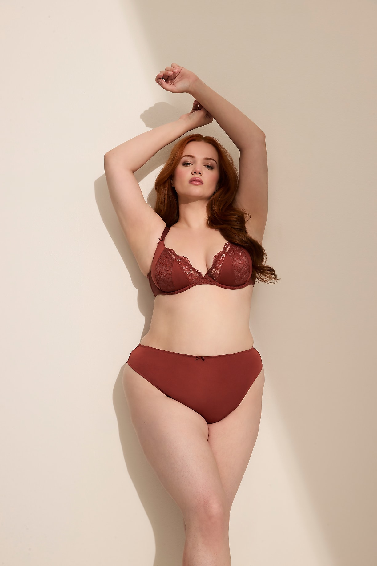 A woman with red hair in a rust-colored bra and underwear poses with her arms raised against a plain, cream-colored wall with a shadow cast on it. Product Image 7