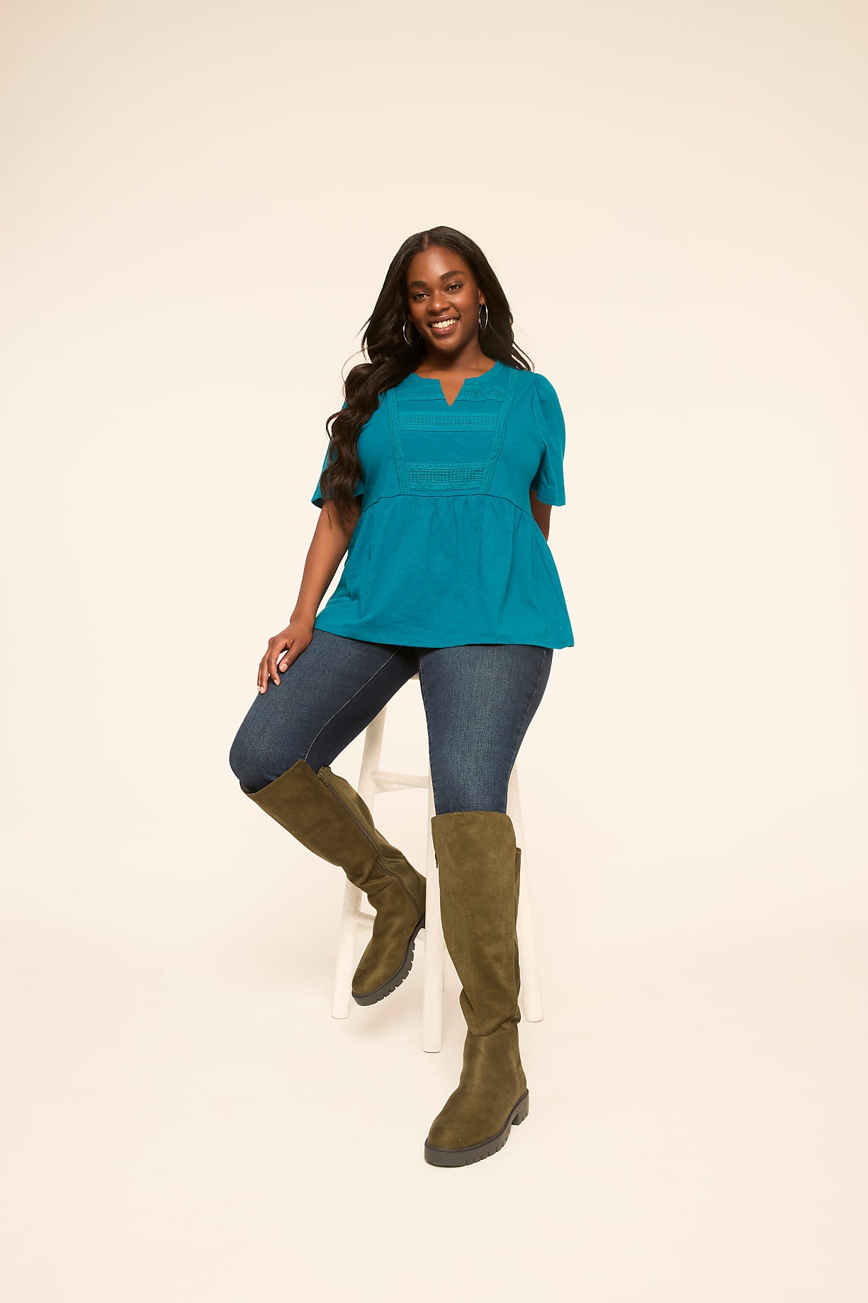 A smiling woman in a teal top, blue jeans, and olive boots sits on a white stool against a plain, light background. Product Image 5