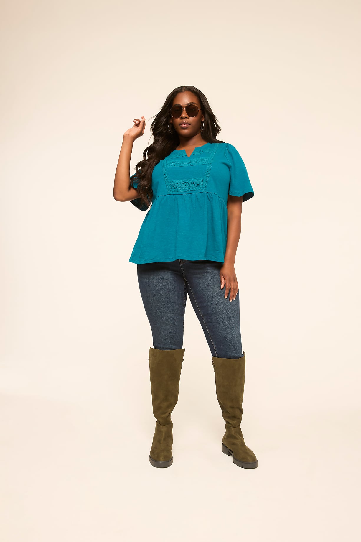 A woman in a teal top, blue jeans, and olive boots stands against a plain cream background, one hand raised to her sunglasses. Product Image 6