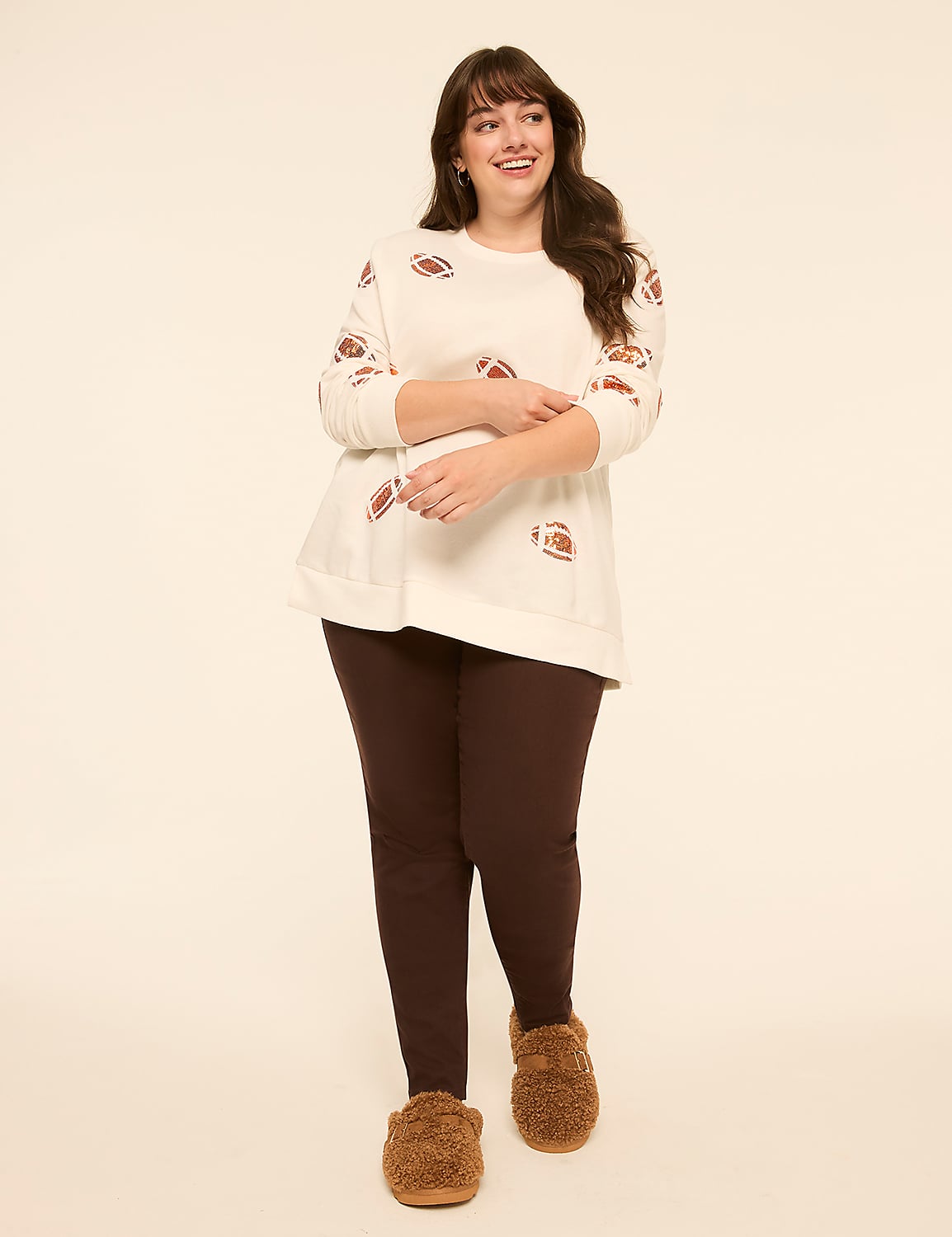 A smiling woman in a cream-colored sweater with football patterns and dark leggings stands on a plain, light-colored background. Product Image 3