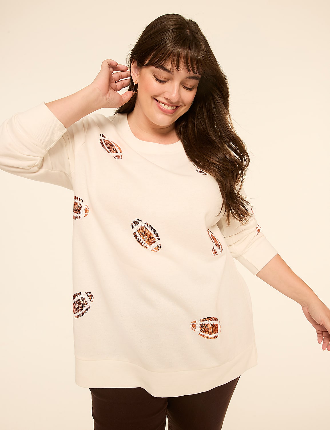 A smiling woman in a cream-colored sweater with football patterns stands against a plain, light-colored background. Product Image 1