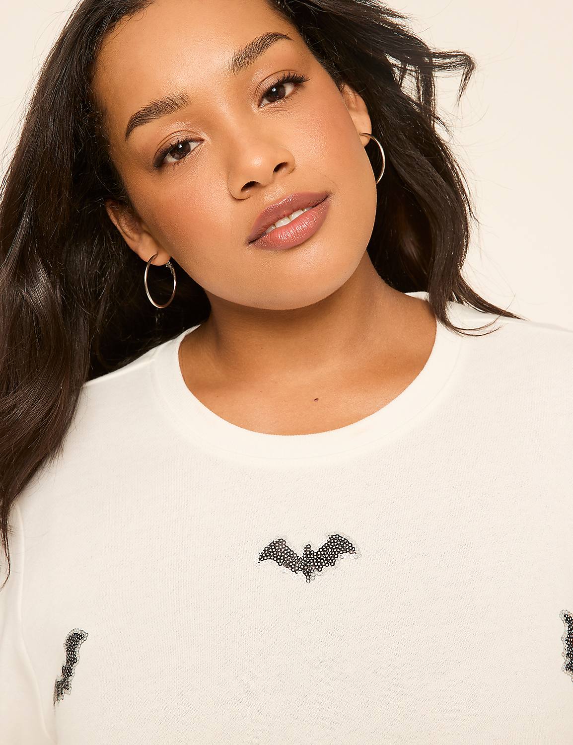 A woman with dark hair and hoop earrings smiles gently in the foreground, wearing a cream-colored sweater adorned with bat designs against a plain, light background. Product Image 4