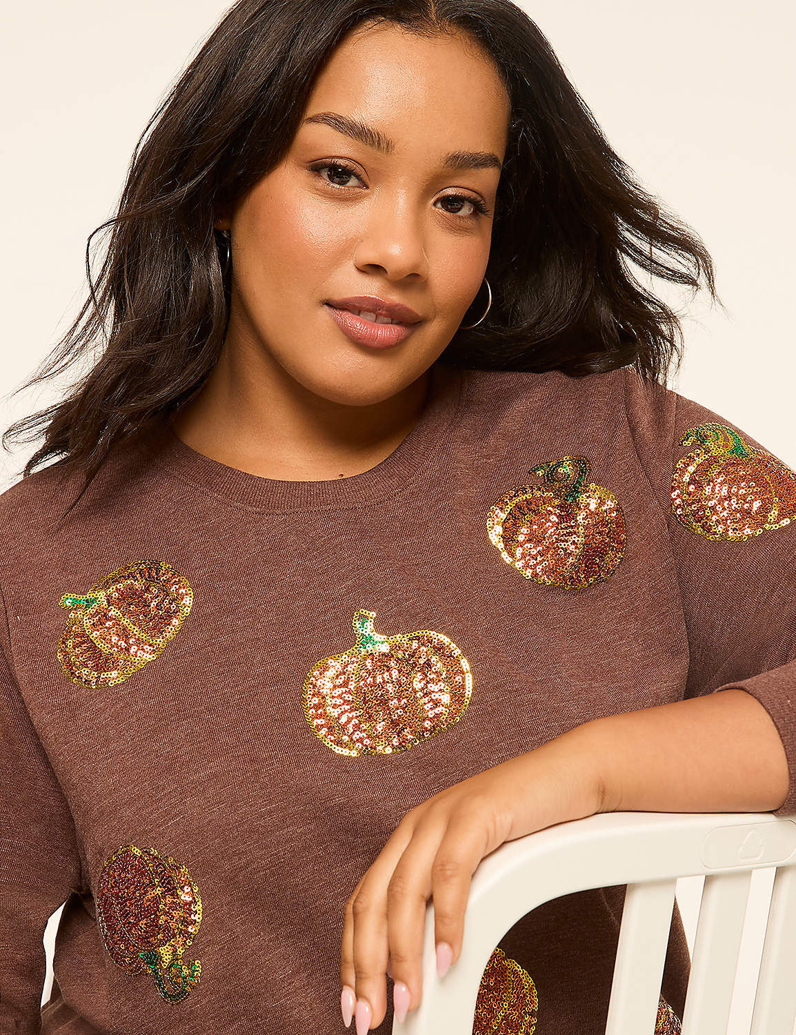 A woman with dark hair and a brown sequined pumpkin sweatshirt leans against a white chair with a plain, light-colored background. Product Image 4