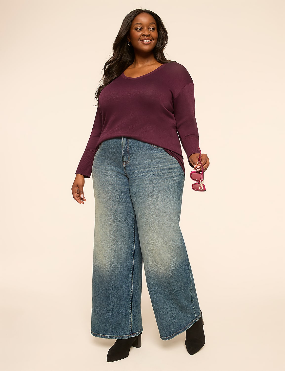 A smiling woman in a plum v-neck sweater and wide-leg jeans stands against a plain, light-colored background. Product Image 3