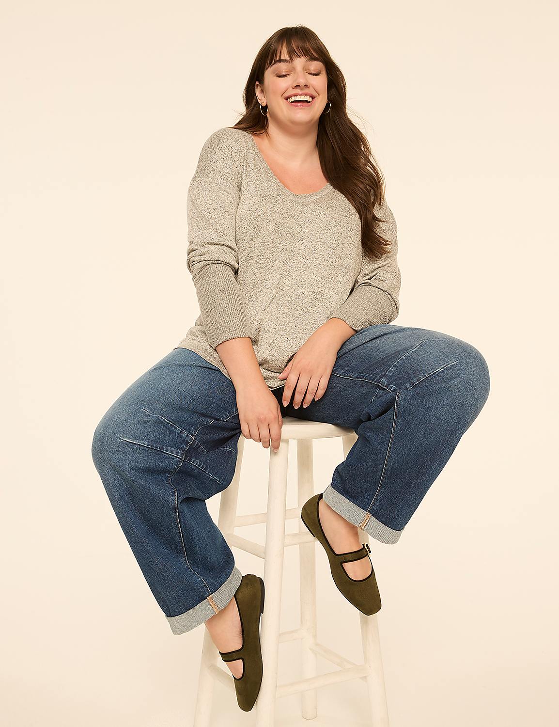 A woman in a grey sweater and baggy jeans laughs while sitting on a white stool against a plain, light-colored background. Product Image 3