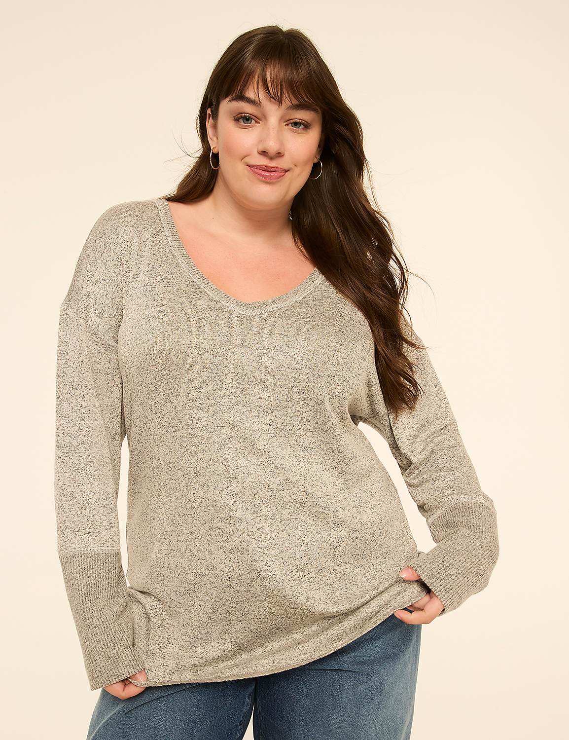 A woman in a gray v-neck sweater and blue jeans smiles against a plain, light-colored background. Product Image 1