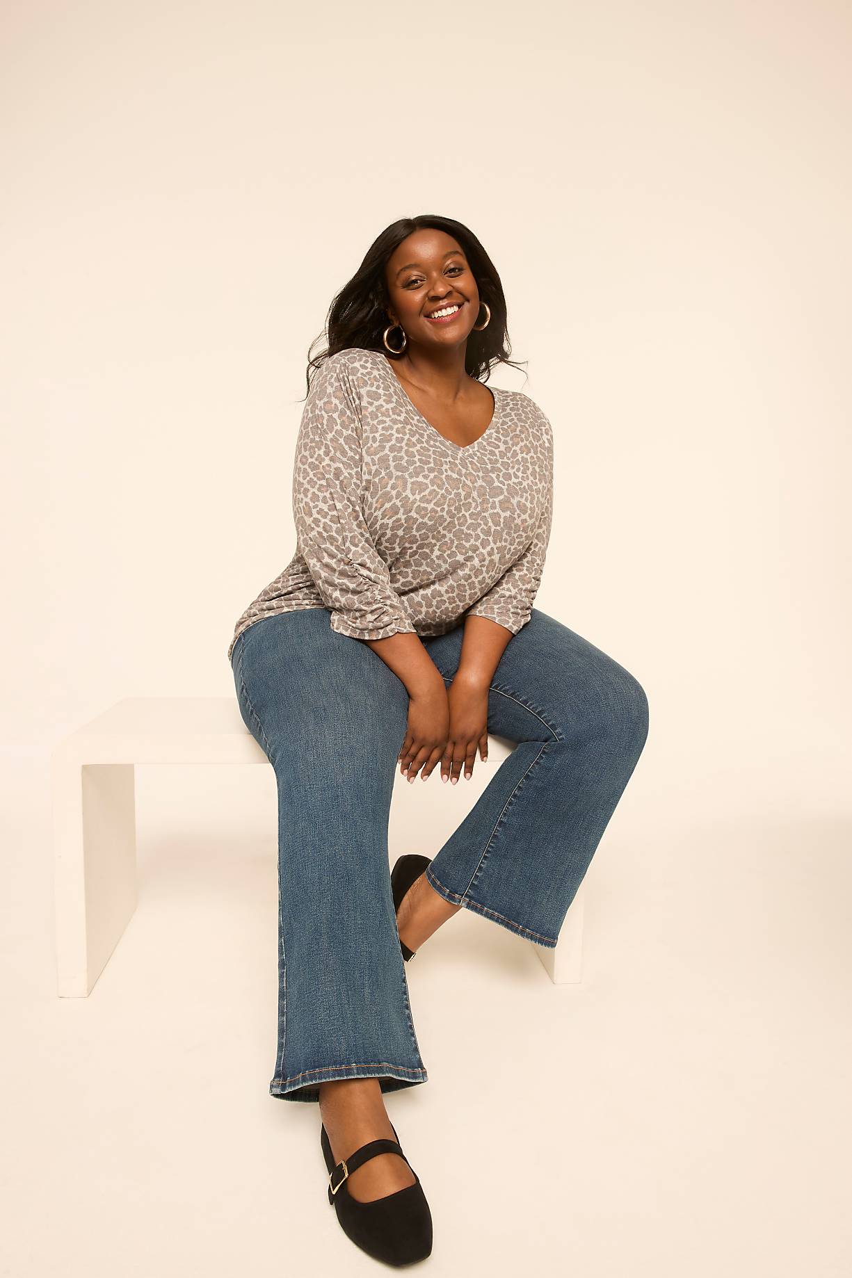 A smiling woman in a leopard print top and blue jeans sits on a white bench against a plain, light background. Product Image 6