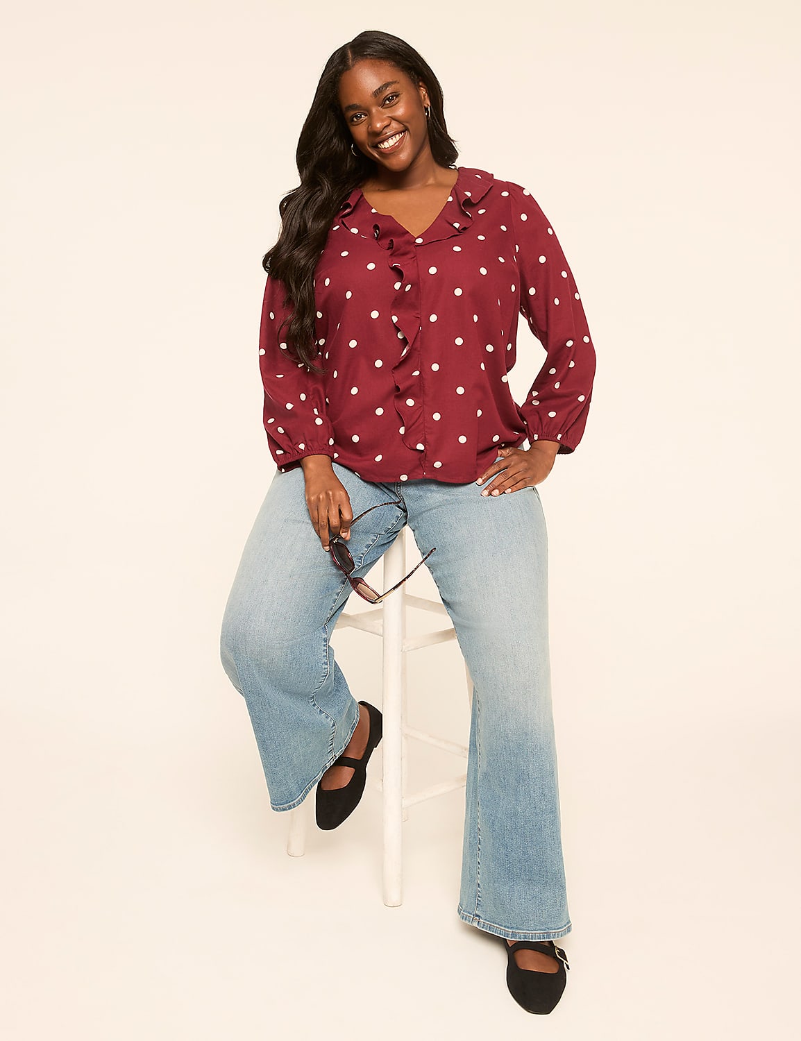 A smiling woman with long dark hair, wearing a polka-dot blouse and flared jeans, sits on a white stool against a plain, light-colored background. Product Image 3