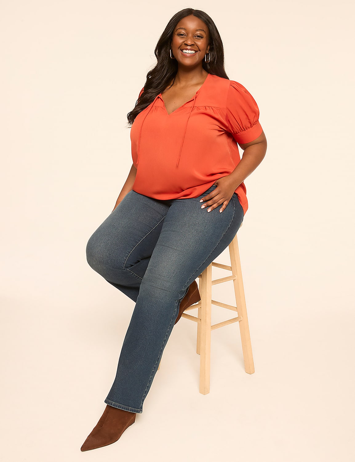 A smiling woman in an orange top and blue jeans sits on a stool against a plain, light-colored background. Product Image 3