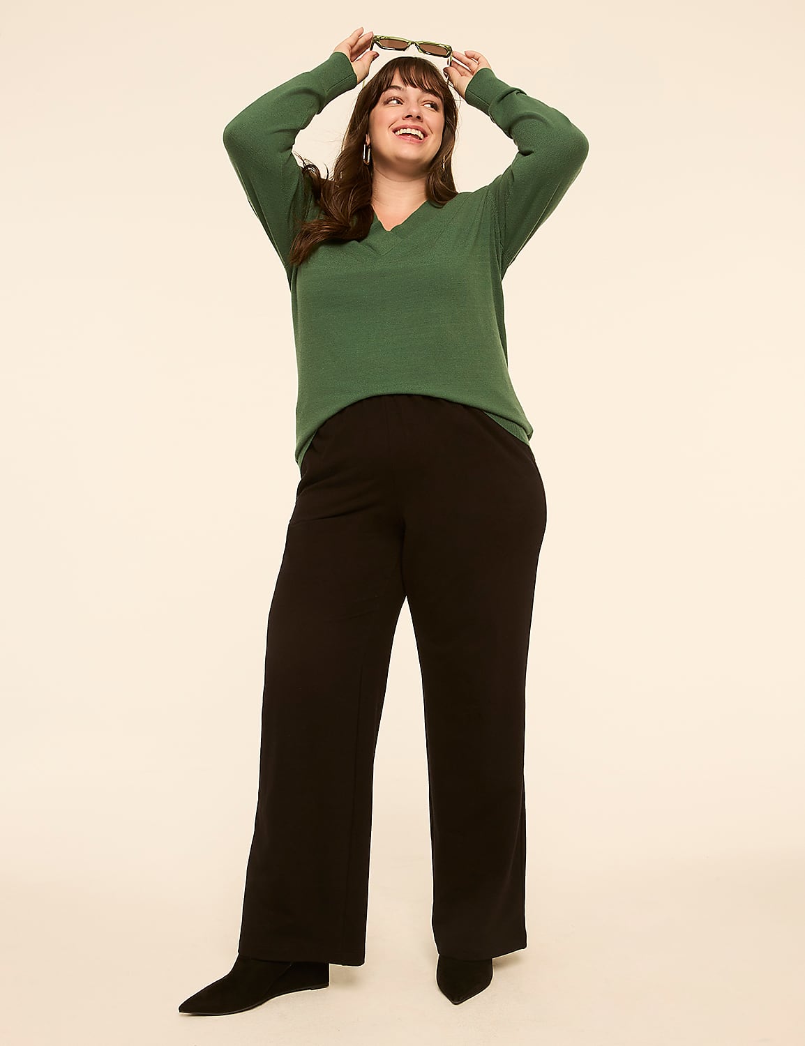A woman in a green sweater and black pants poses with sunglasses above her head against a plain, light-colored background. Product Image 3