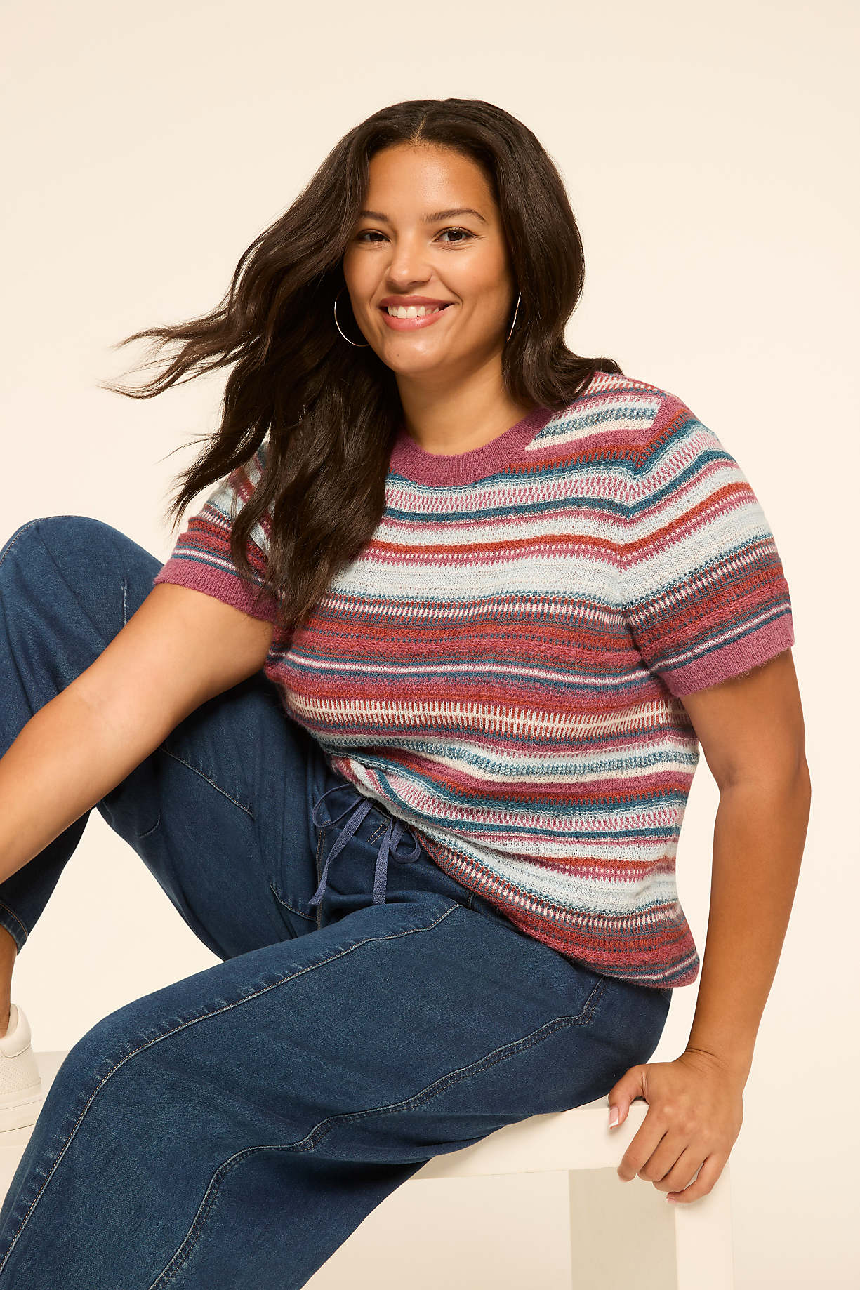 A smiling woman in a striped sweater and jeans sits in front of a plain, light-colored background. Product Image 5