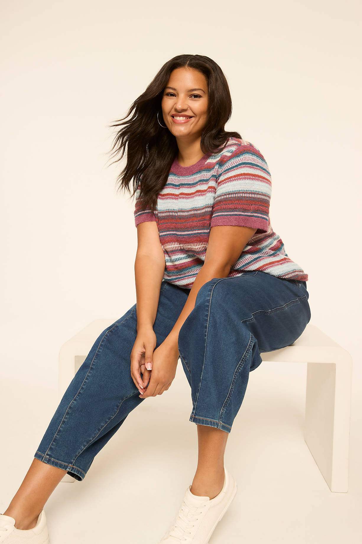 A smiling woman in a striped shirt and jeans sits on a white bench against a plain, light background. Product Image 6