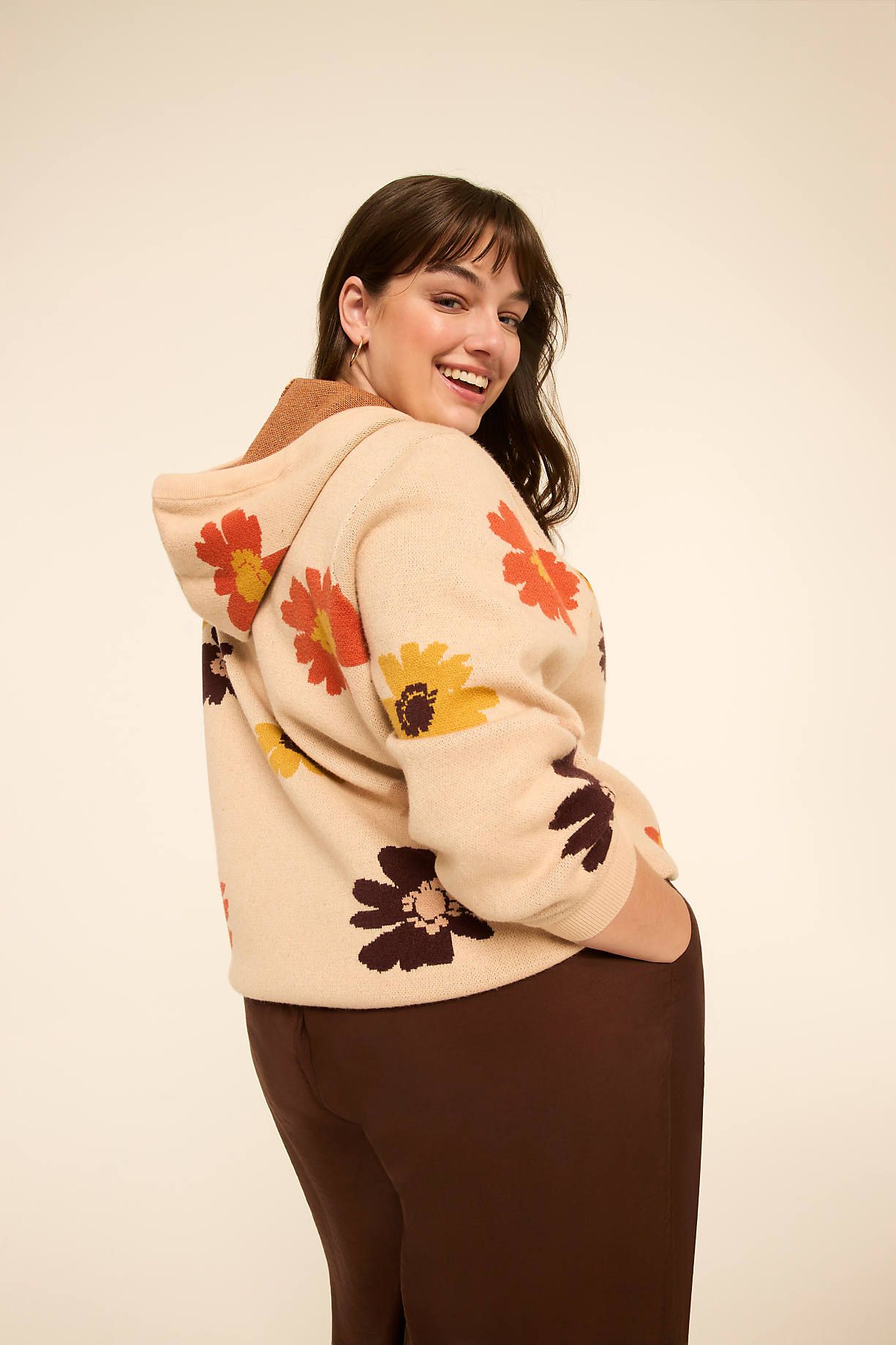 A smiling woman in a cream-colored, floral-patterned hoodie and brown pants stands against a plain, light-colored background. Product Image 6