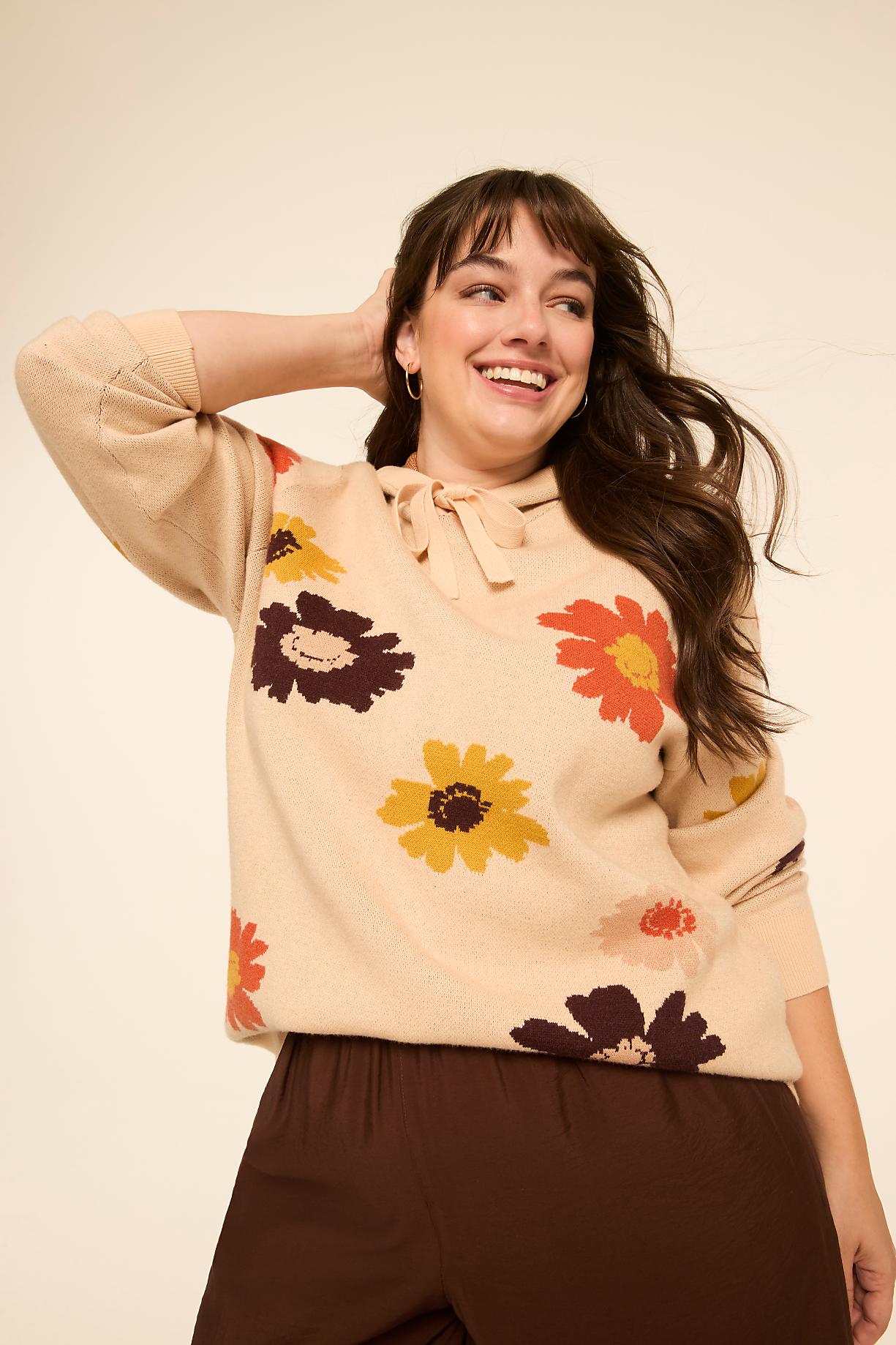A smiling woman in a beige sweater with large floral patterns stands against a plain, light background. Product Image 7