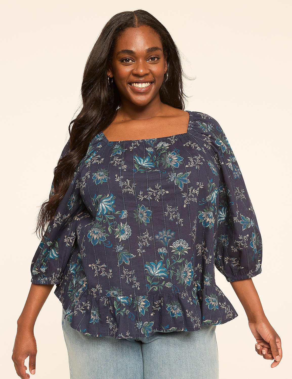 A smiling woman with long dark hair wears a dark blue floral print blouse with a ruffled hem, standing against a plain, light-colored background. Product Image 1