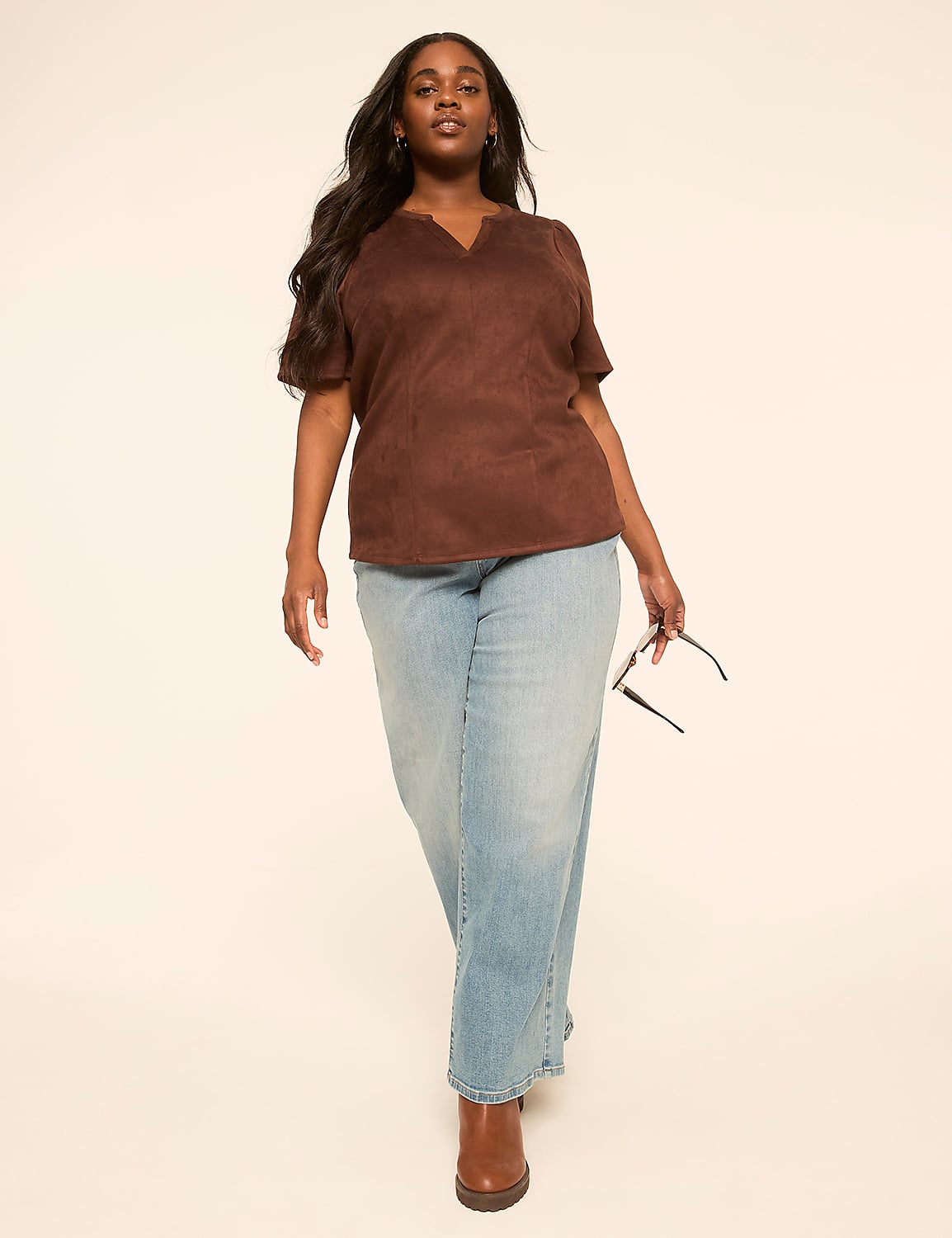 A woman in a brown top and light blue jeans walks forward against a plain, light background, holding sunglasses in her right hand. Product Image 3