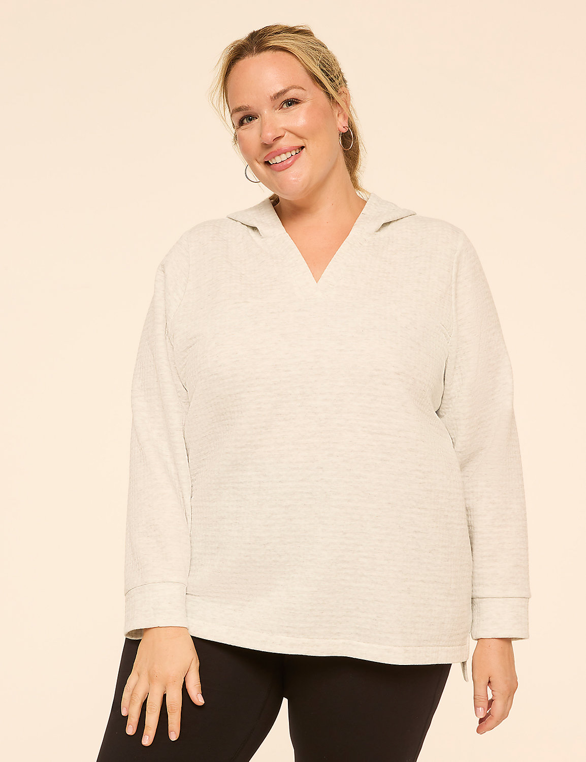 A smiling woman in a light grey hoodie and black leggings stands against a plain, light peach background. Product Image 1