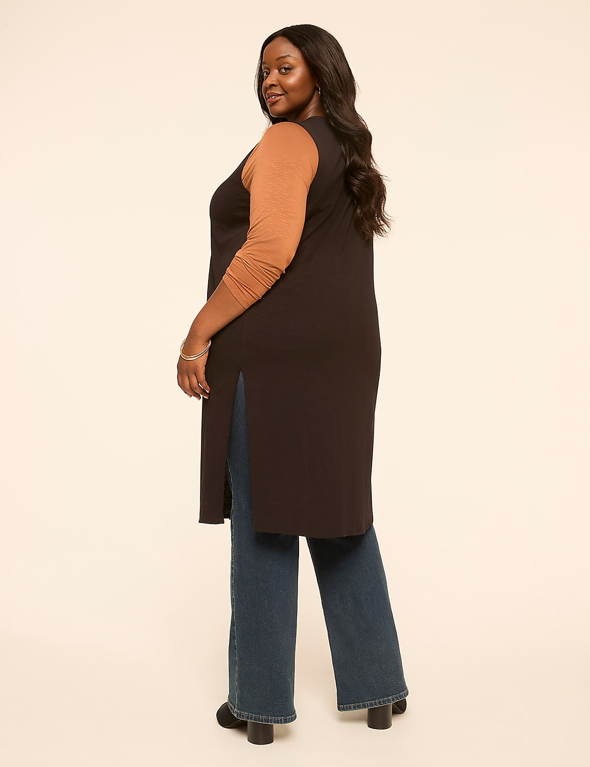 A woman in a long dark vest over a tan long-sleeved shirt and blue jeans stands against a plain, light-colored background. Product Image 2
