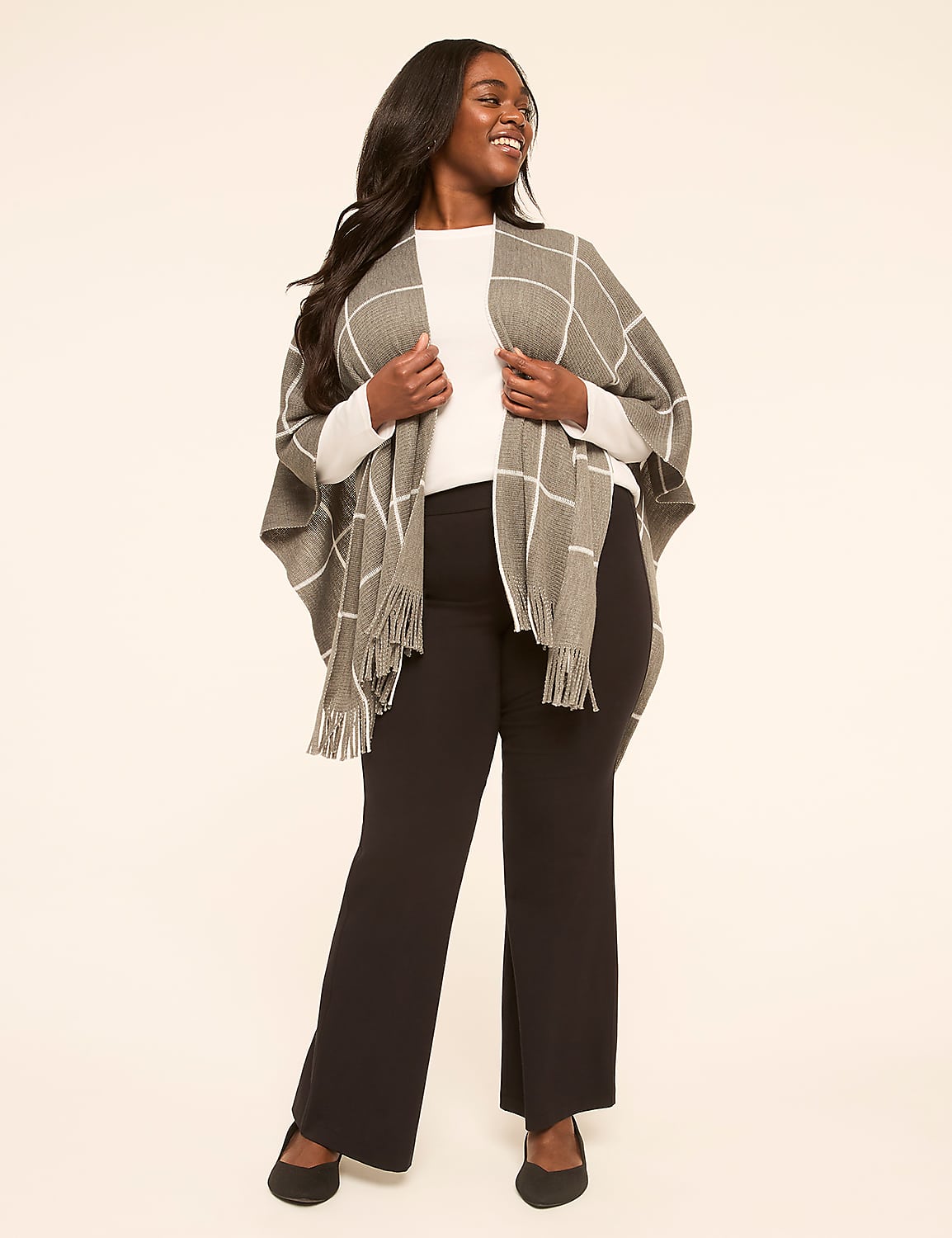 A smiling woman in a white top, black pants, and a gray checkered poncho with fringe stands against a plain, light-colored background. Product Image 3