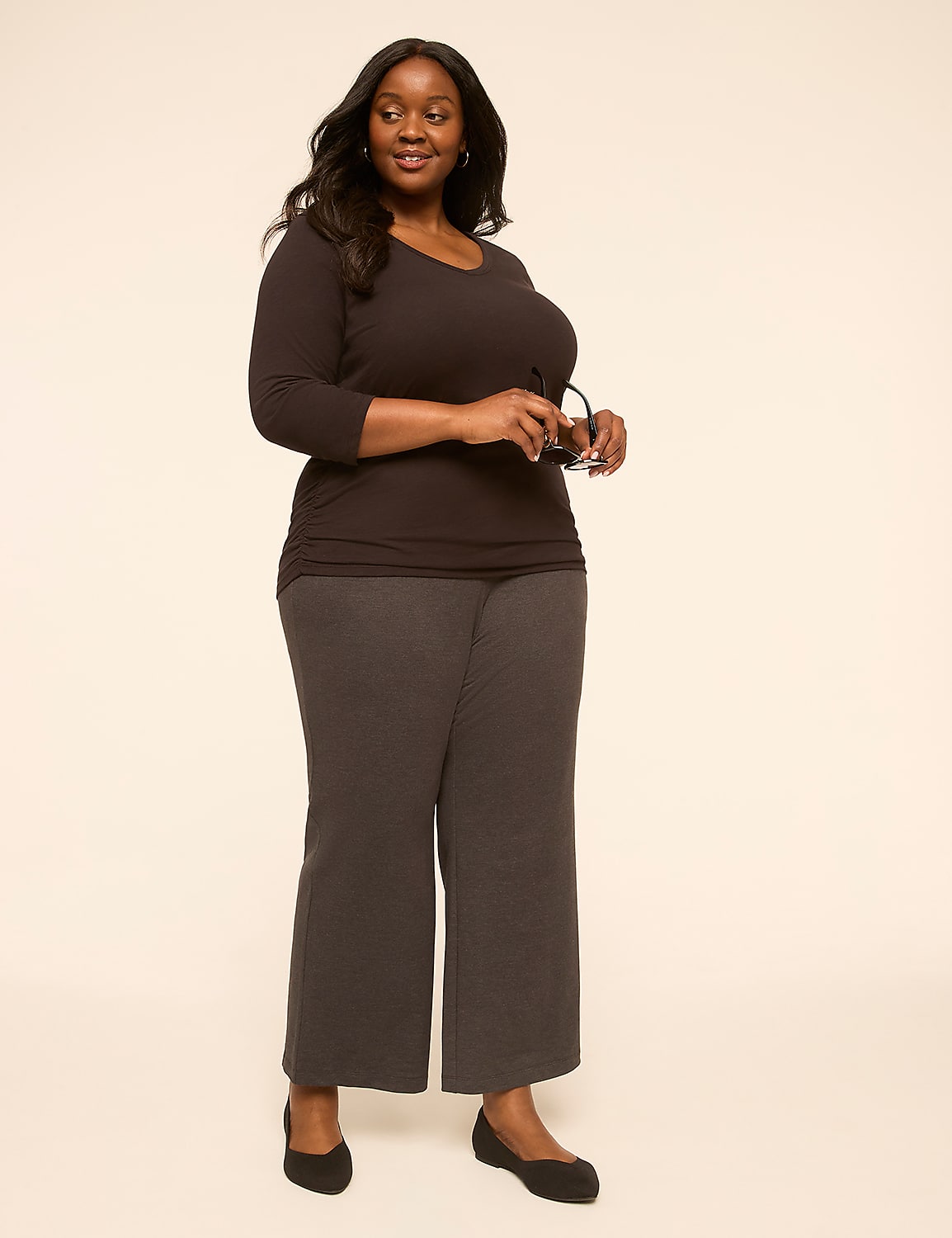A woman in a dark top and gray pants stands against a plain, light-colored background, holding glasses in her right hand. Product Image 3