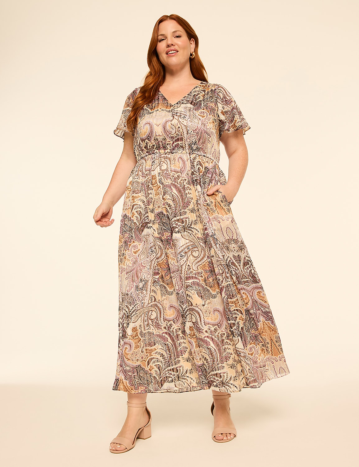 A woman in a flowing paisley dress with flutter sleeves stands with her hands in her pockets against a plain, light-colored background. Product Image 1