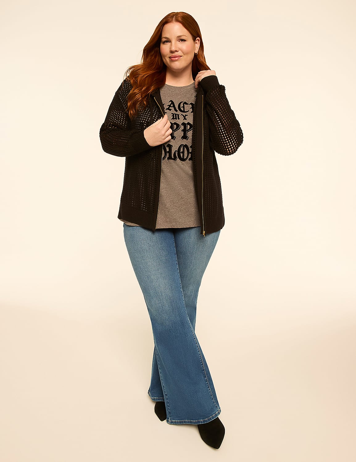A woman with red hair stands in the foreground wearing a black zip-up jacket over a graphic t-shirt and flared jeans, against a plain, light-colored background. Product Image 3