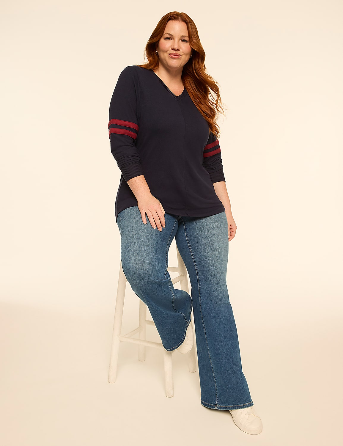 A woman with auburn hair sits on a white stool in the foreground, wearing a navy v-neck shirt with red stripes on the sleeves and flared blue jeans, against a plain, light-colored background. Product Image 3
