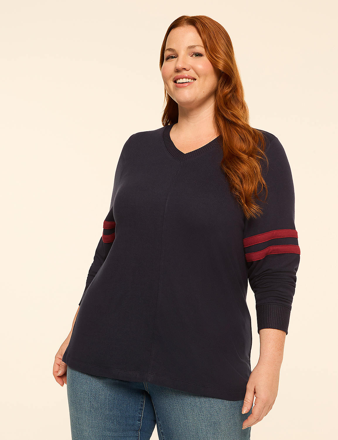 A smiling woman with red hair in a navy v-neck sweater with burgundy stripes on the sleeves stands against a plain cream background. Product Image 1