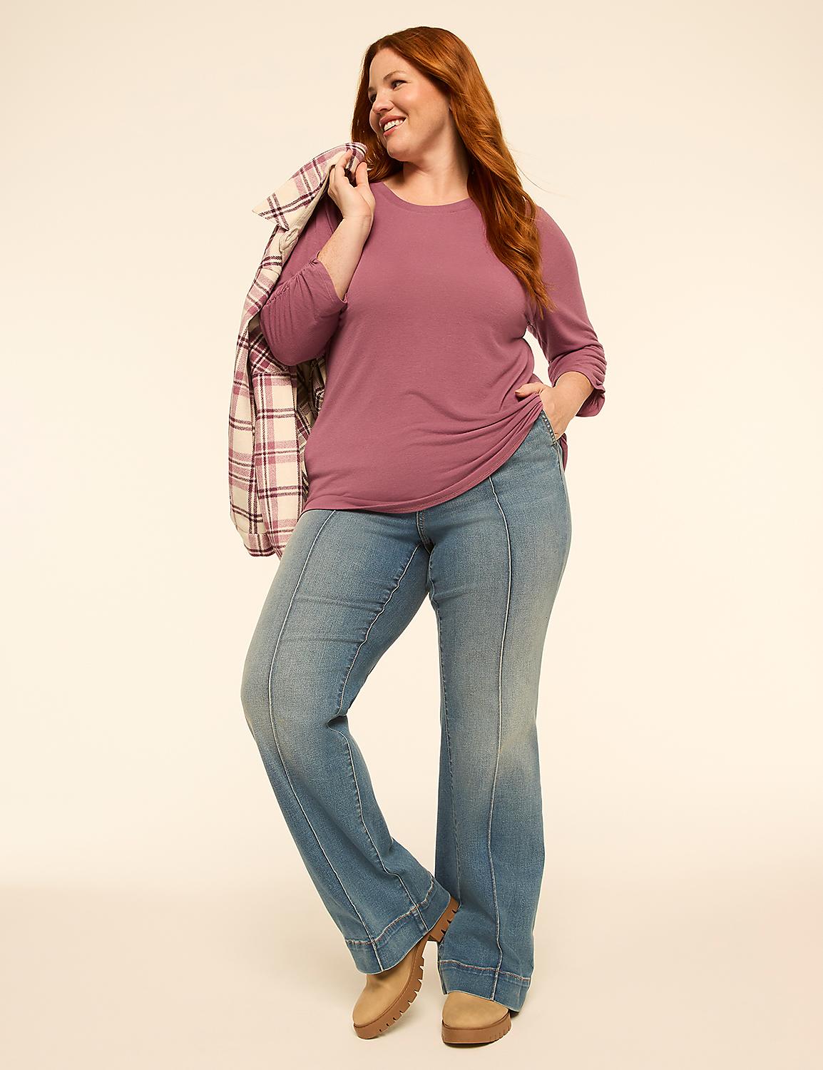A smiling woman with red hair stands in the foreground wearing a mauve long-sleeved shirt, flared jeans, and tan boots, with a plaid jacket draped over her shoulder, against a plain, light-colored background. Product Image 3
