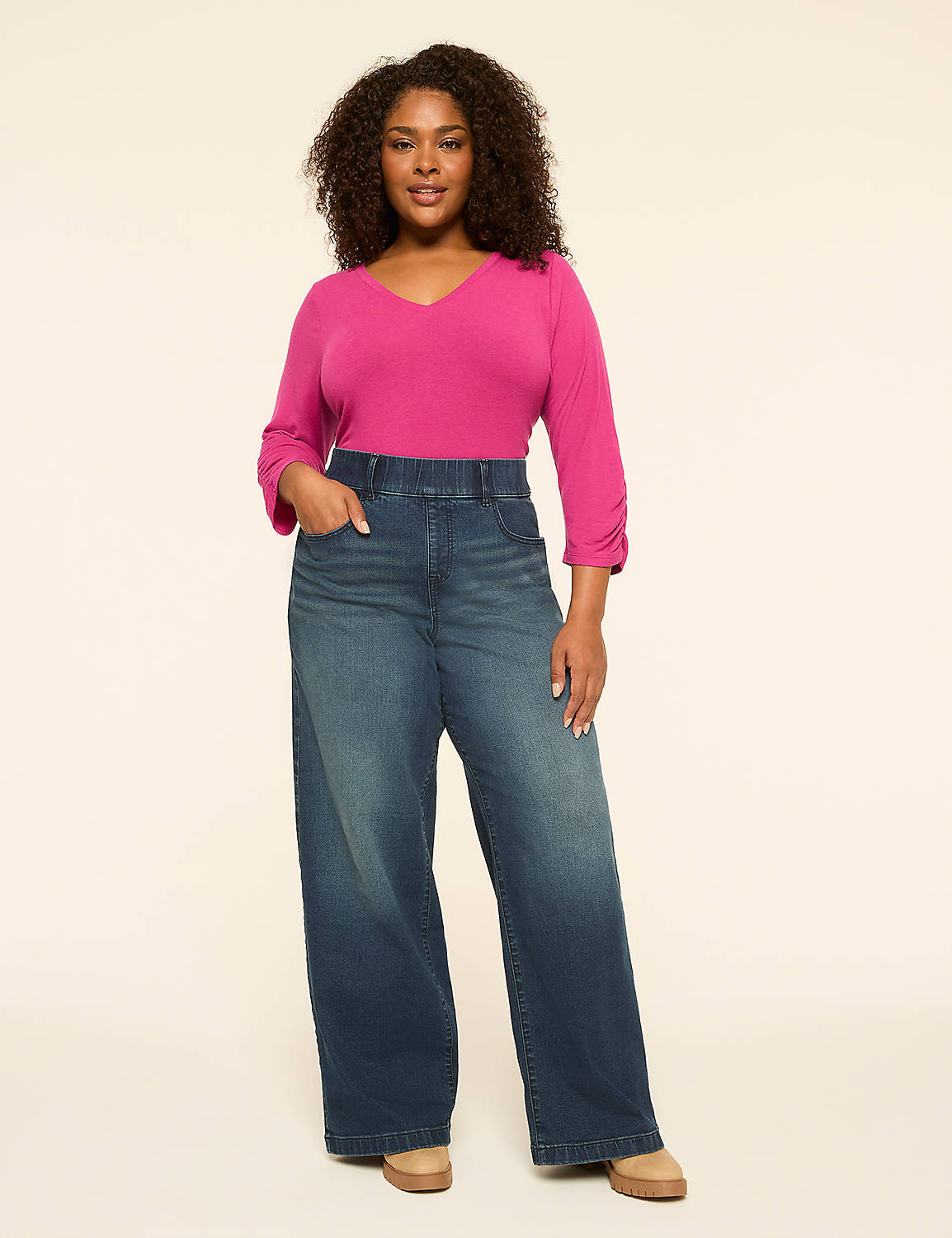 A woman with curly dark hair stands in the foreground wearing a bright pink v-neck shirt and wide-leg denim jeans against a plain, light-colored background. Product Image 3