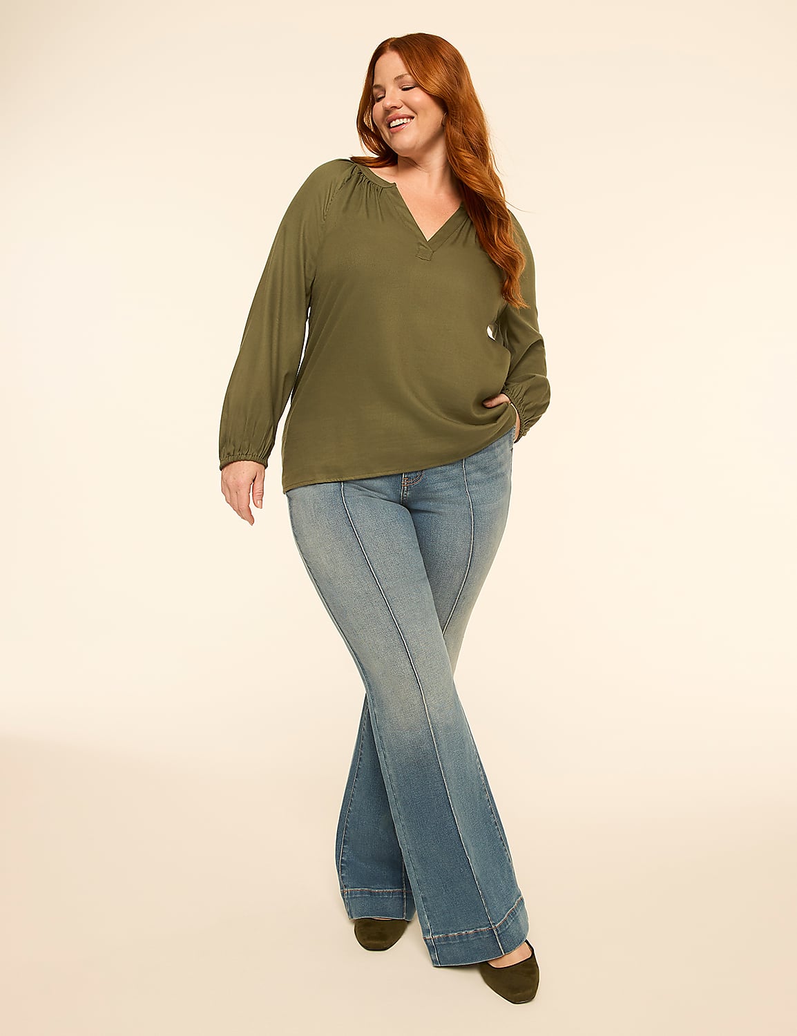 A smiling woman with auburn hair poses in a green peasant blouse and flared jeans against a plain, light-colored backdrop. Product Image 3