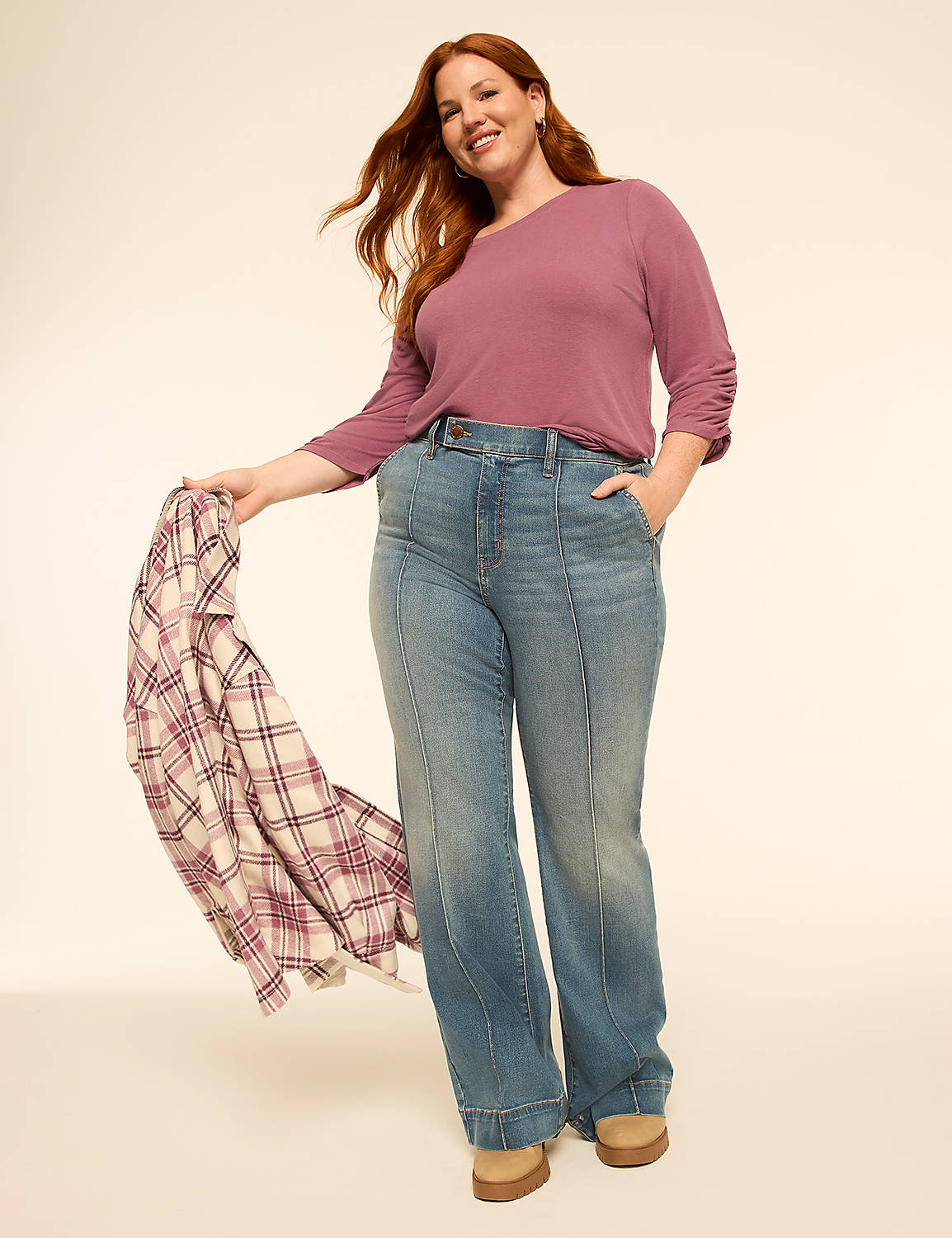 A smiling woman with red hair in a purple shirt and wide-leg jeans stands against a plain, light-colored background, holding a plaid shirt. Product Image 3