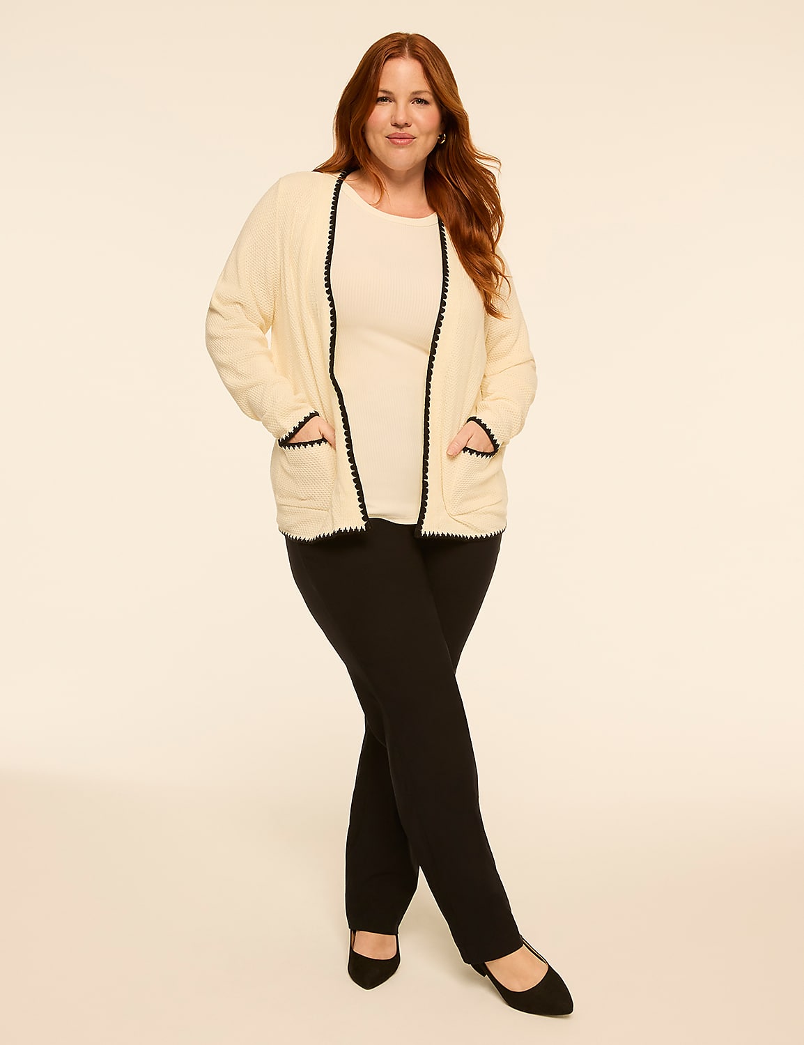 A woman with red hair stands with her legs crossed in the foreground, wearing a cream cardigan over a cream top and black pants, against a plain cream background. Product Image 3