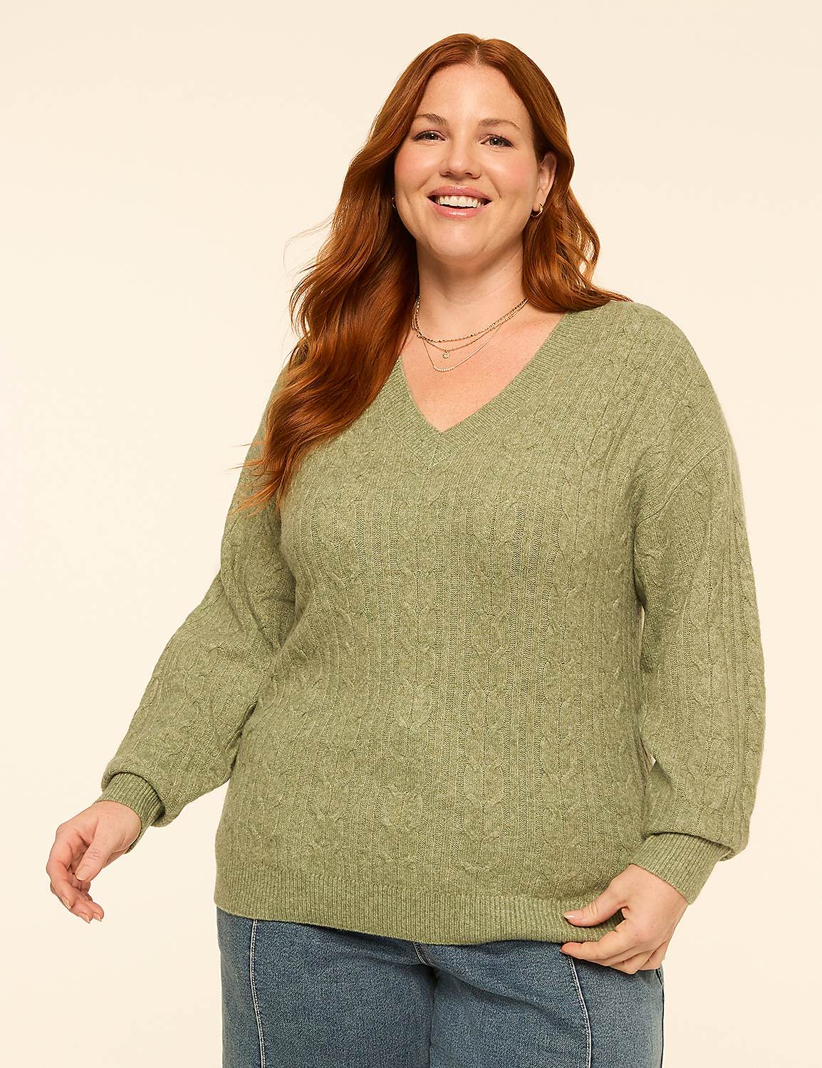 A smiling woman with red hair wears a green cable-knit sweater and blue jeans against a plain, light-colored background. Product Image 1