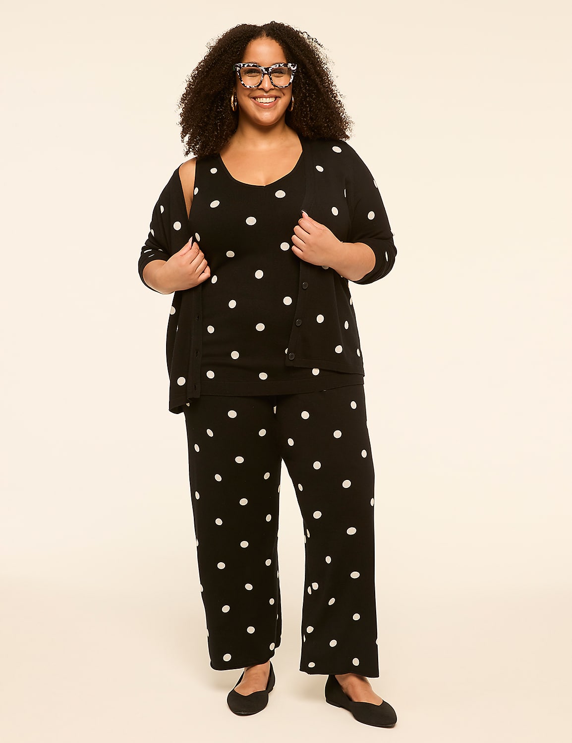 A smiling woman with curly hair and large glasses stands in the foreground wearing a black polka dot pajama set and cardigan against a plain, light-colored background. Product Image 3