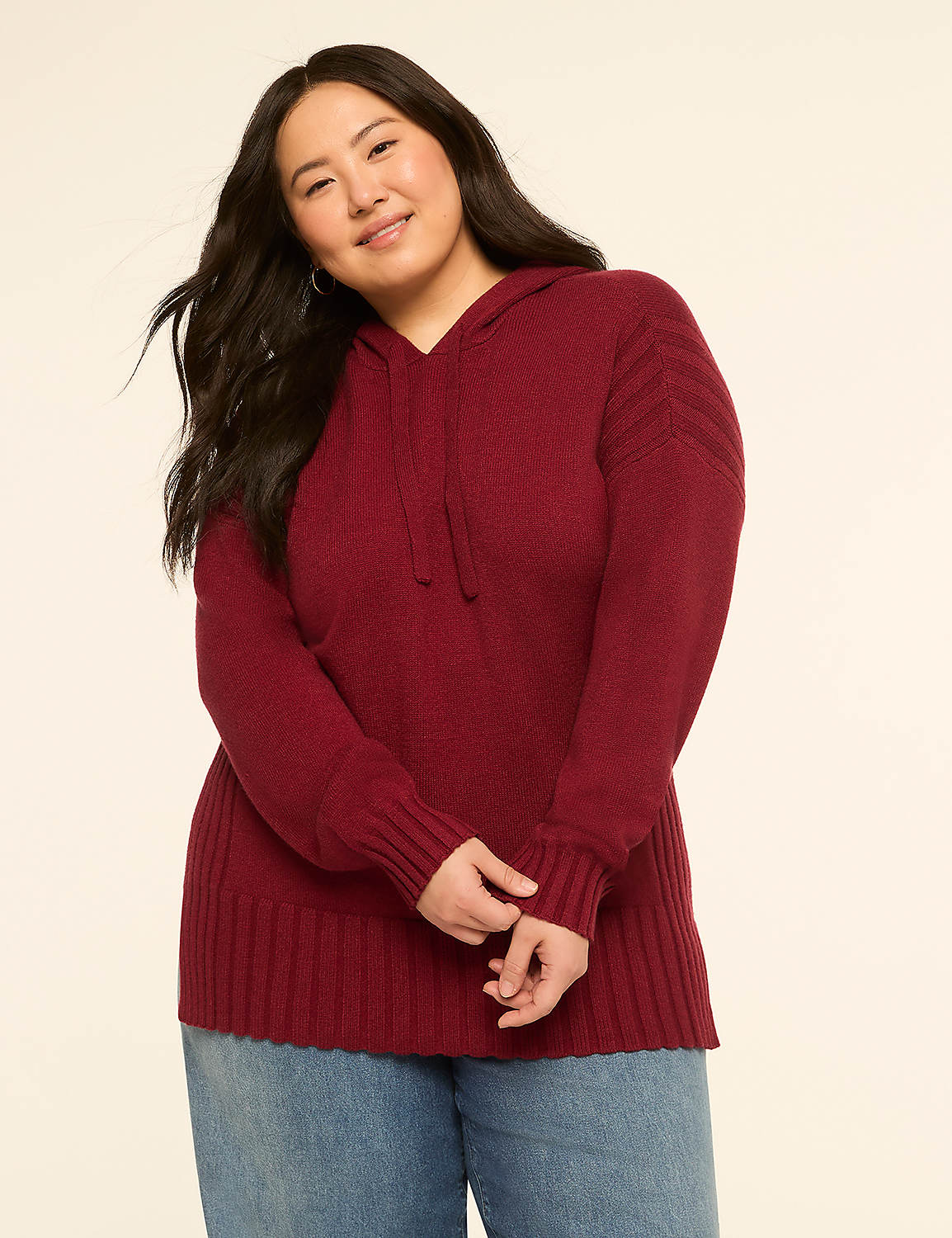 A smiling woman in a maroon hooded sweater and blue jeans stands against a plain cream background. Product Image 1