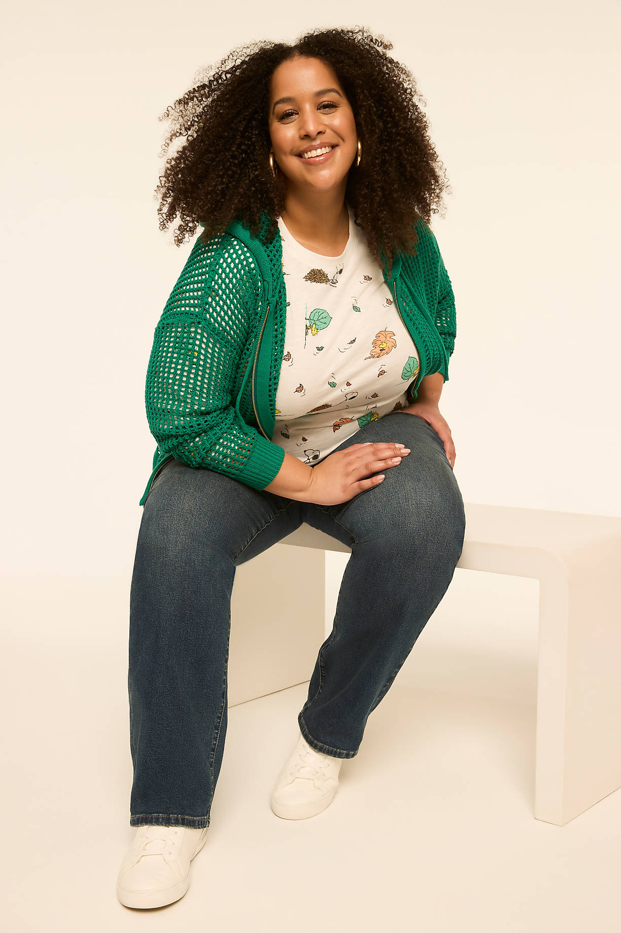 A smiling woman with curly hair, wearing a green cardigan over a graphic t-shirt and jeans, sits on a white bench against a plain, light-colored background. Product Image 6