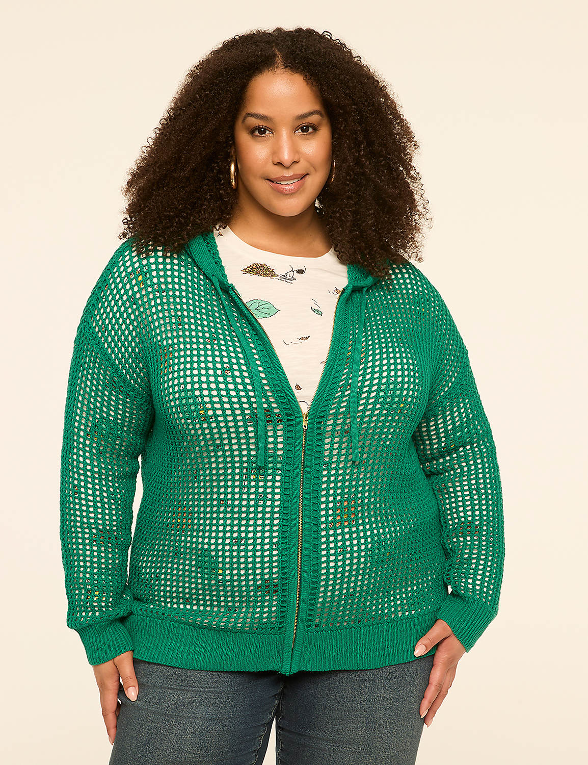 A smiling woman with curly hair wears a green hooded cardigan over a patterned shirt and jeans, standing against a plain, light-colored background. Product Image 1