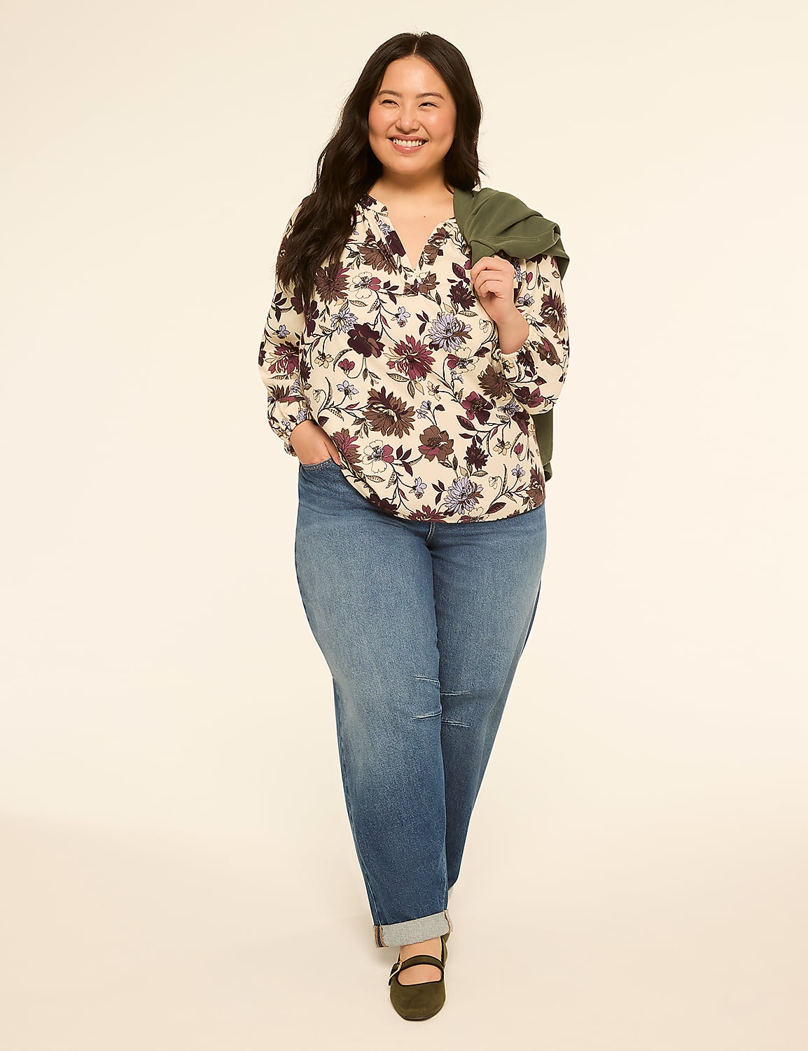 A smiling woman in a floral blouse and jeans stands against a plain, light background with a green jacket draped over her shoulder. Product Image 3