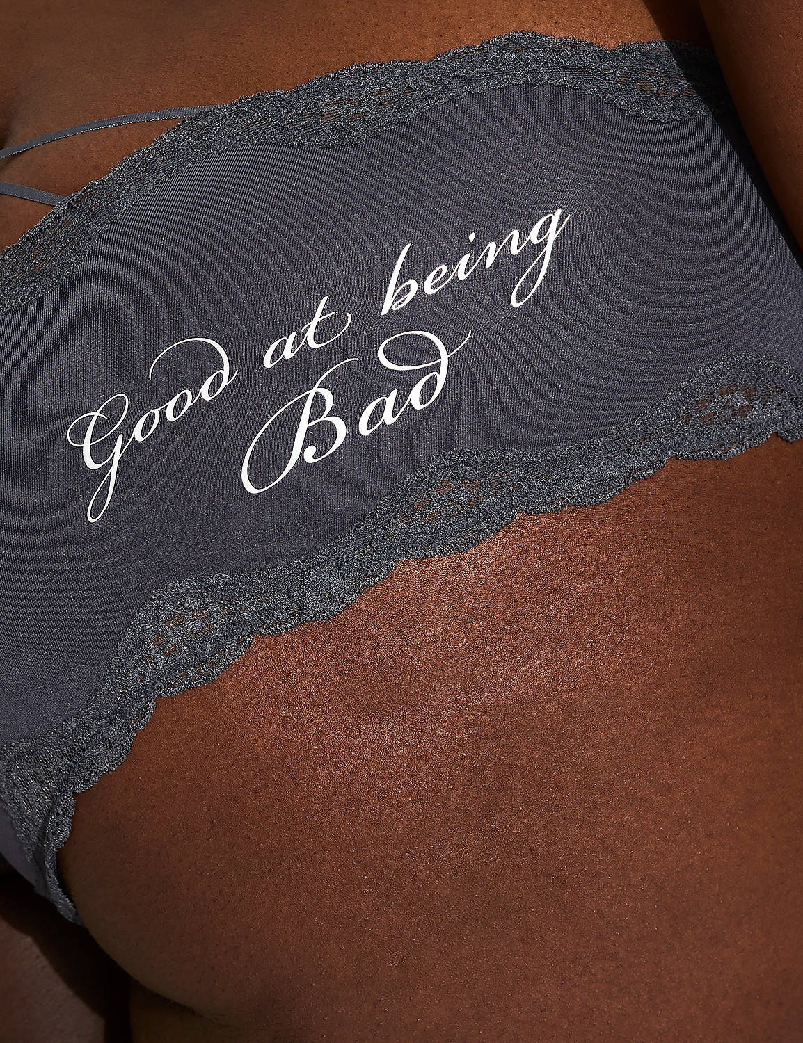 A close-up of dark skin wearing a grey strapless bra with white cursive text reading "Good at being Bad" and lace trim, against a blurred background. Product Image 5