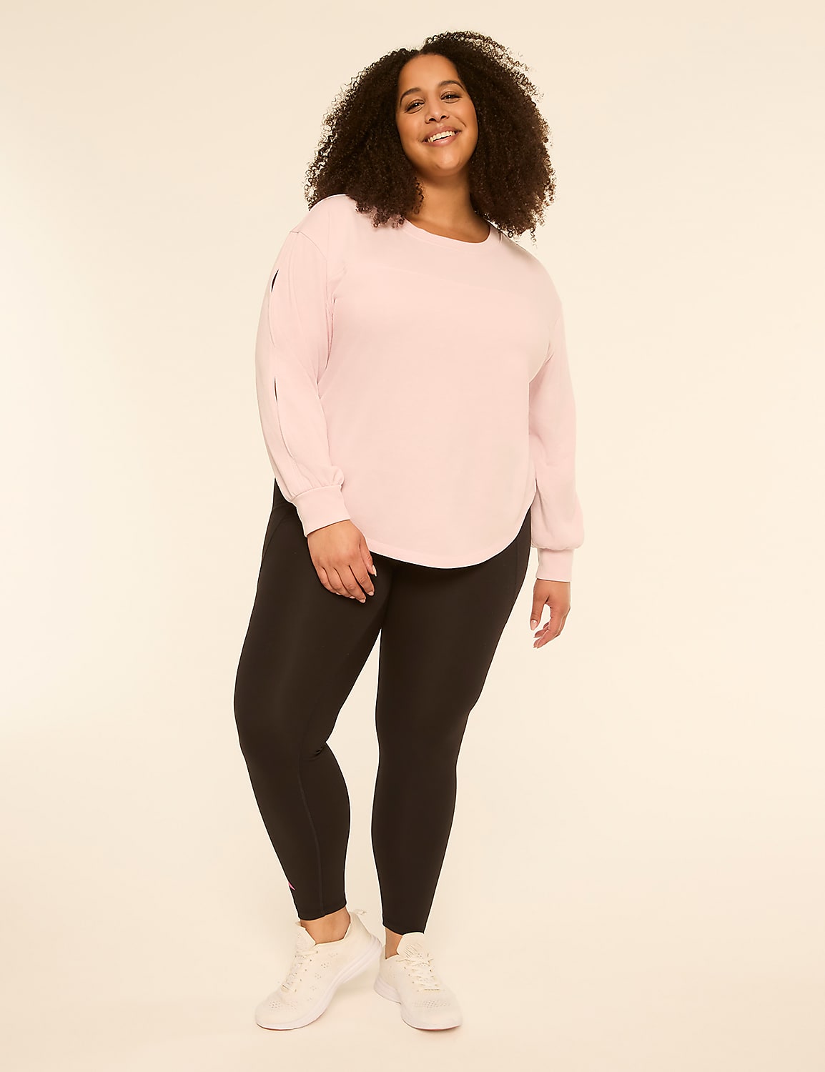 A smiling woman with curly hair stands in the foreground wearing a pink top and black leggings against a plain, light-colored background. Product Image 3