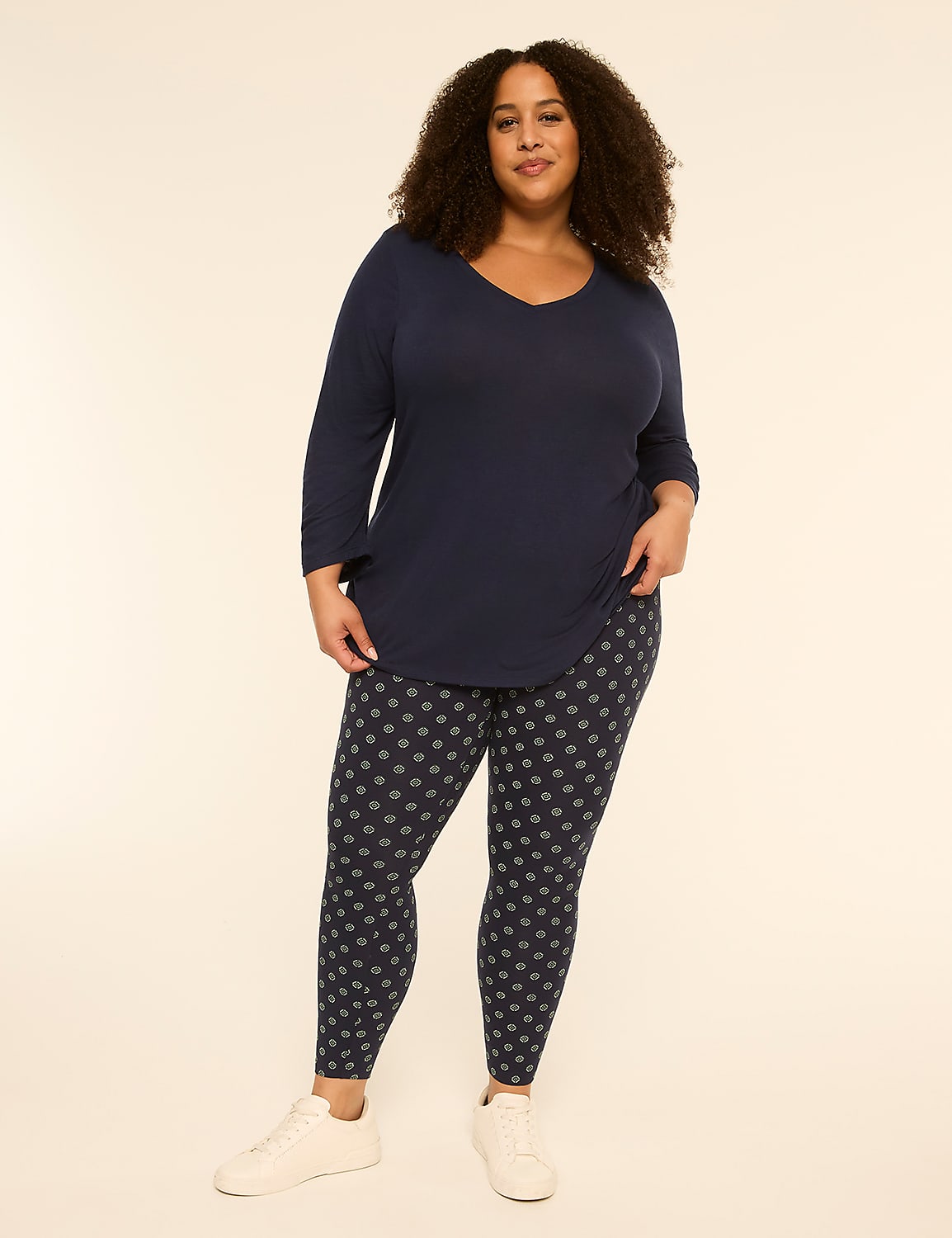 A woman with curly hair stands in the foreground wearing a navy v-neck shirt and patterned leggings, against a plain, light-colored background. Product Image 3
