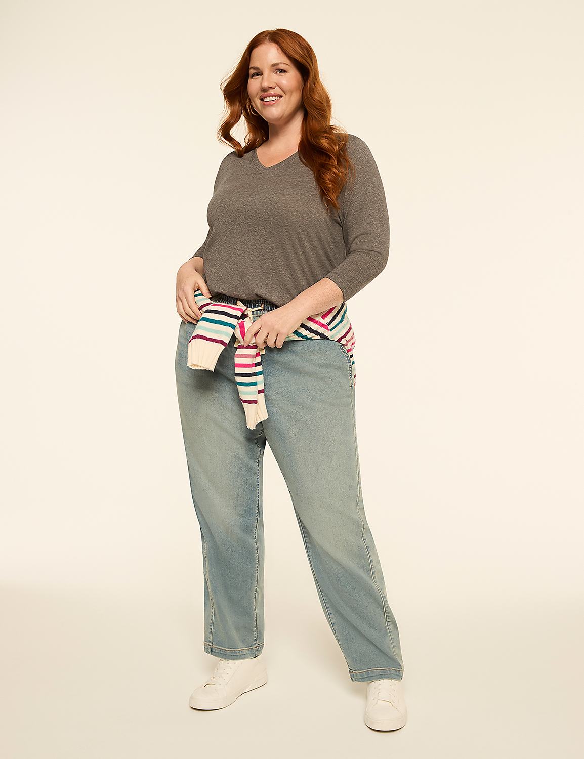 A smiling woman with red hair stands in the foreground wearing a gray v-neck shirt, light wash jeans, and a striped sweater tied around her waist, against a plain, light-colored background. Product Image 3