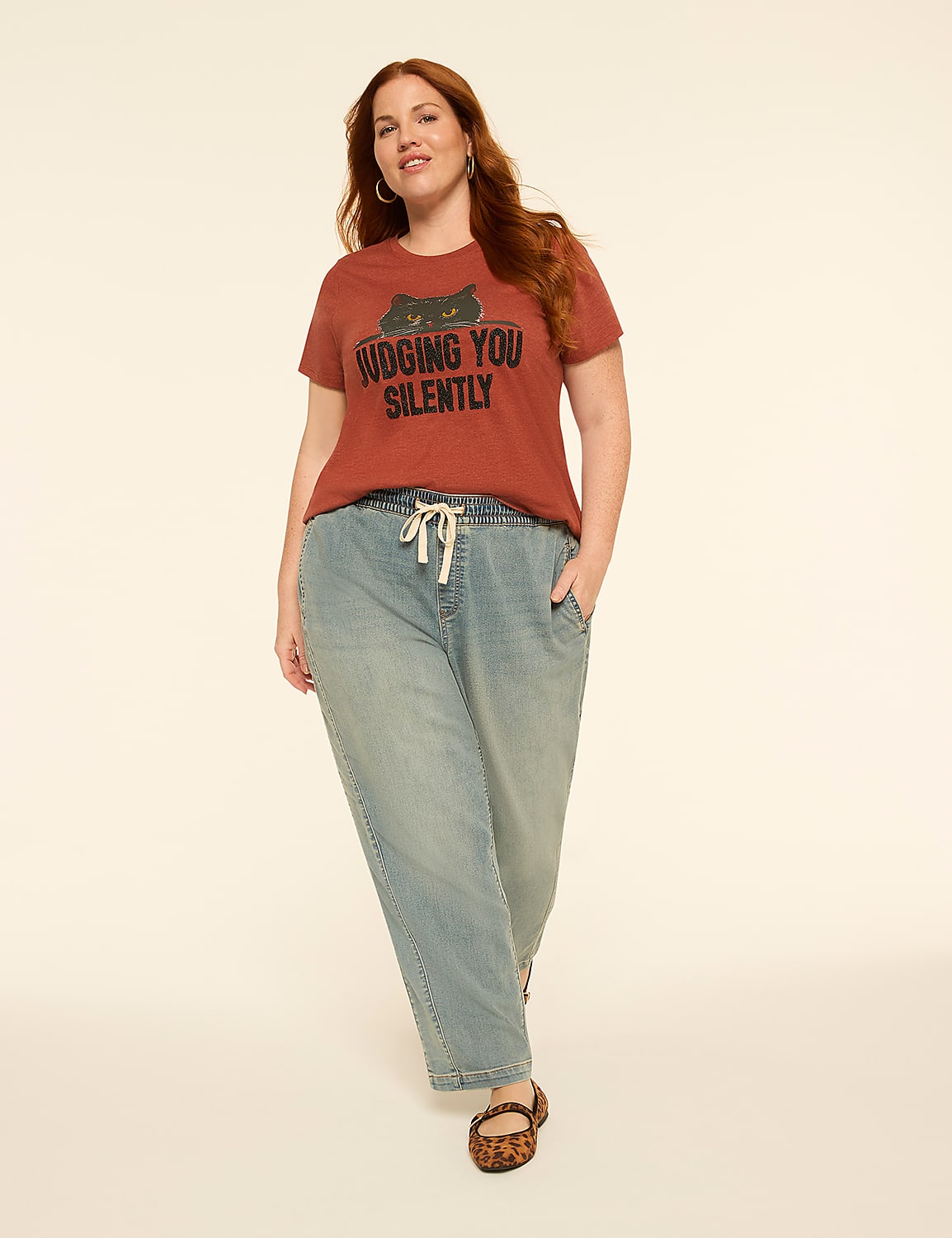 A woman with auburn hair stands in the foreground wearing a rust-colored t-shirt with a cat graphic and the words "JUDGING YOU SILENTLY," paired with light-wash denim pants, against a plain, light-colored background. Product Image 3