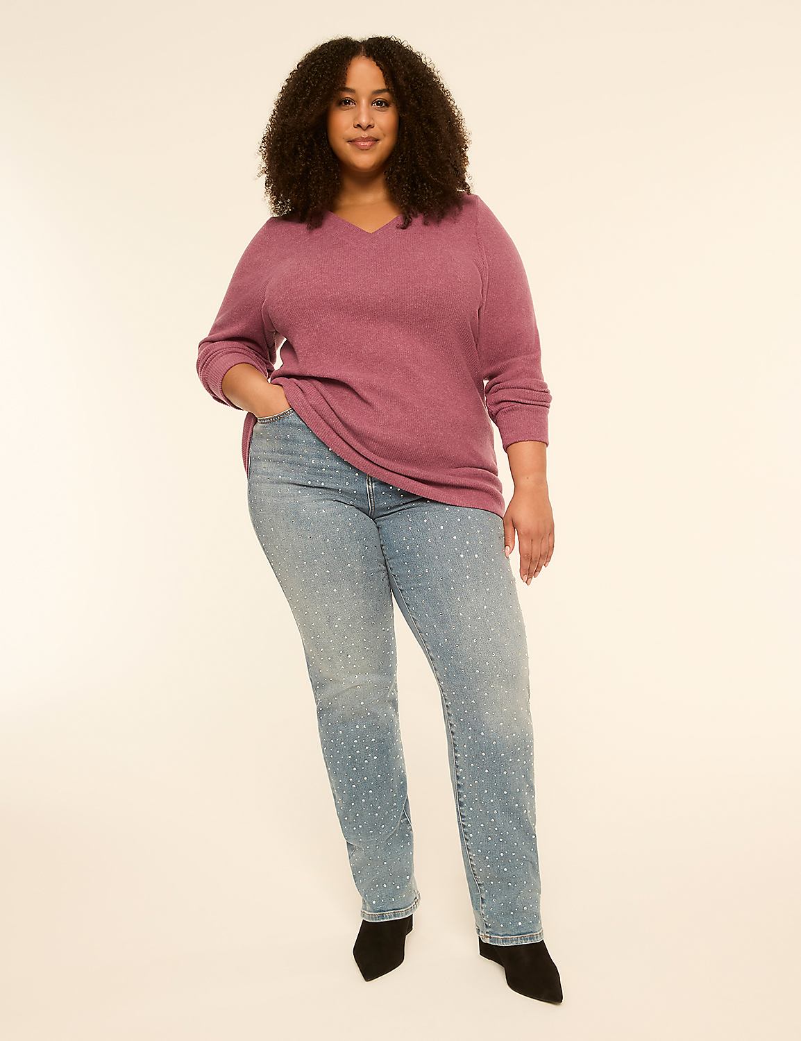 A woman with curly hair stands in the foreground wearing a pink v-neck sweater and embellished jeans against a plain, light-colored background. Product Image 3